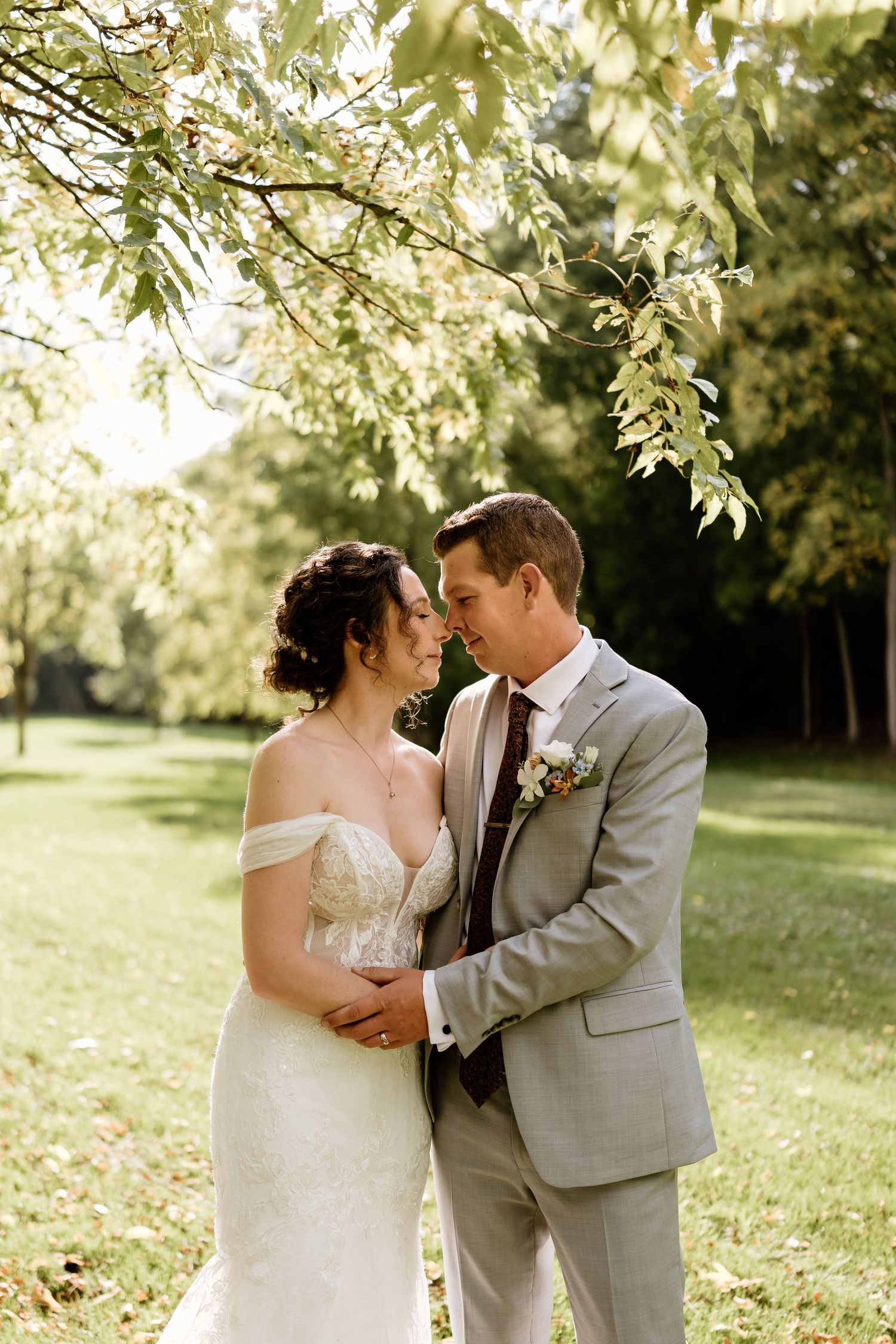 An intimate wedding portrait beneath sunlit tree branches in a natural outdoor setting.