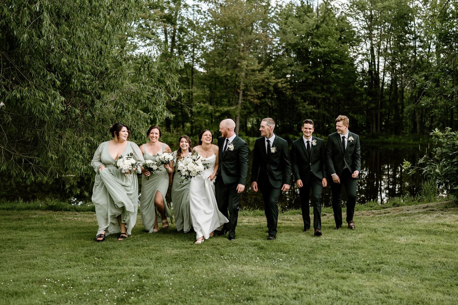 Wedding party walks and laughs together on green lawn with trees in background.
