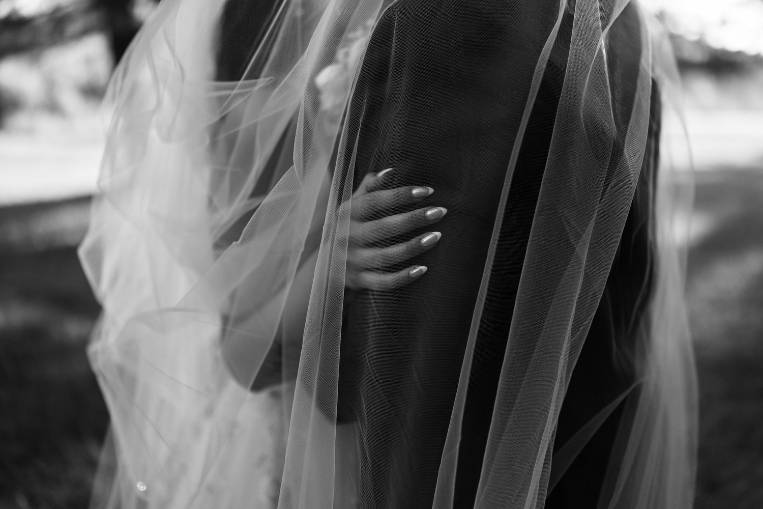 Black and white wedding photo of bride's hand with manicured nails resting on groom's suit under flowing veil.