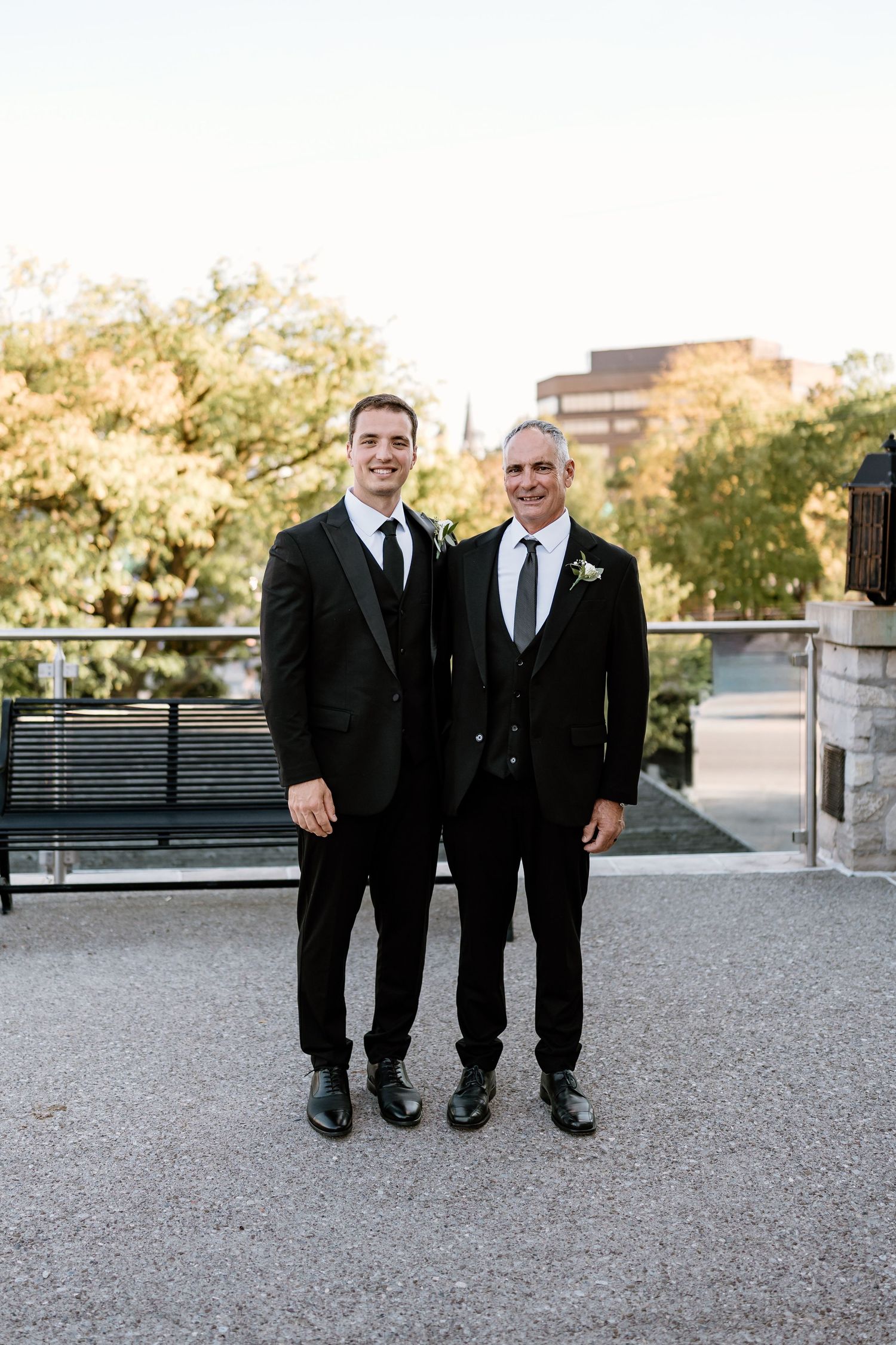 Three groomsmen in black suits and ties stand together on gravel path at outdoor venue.