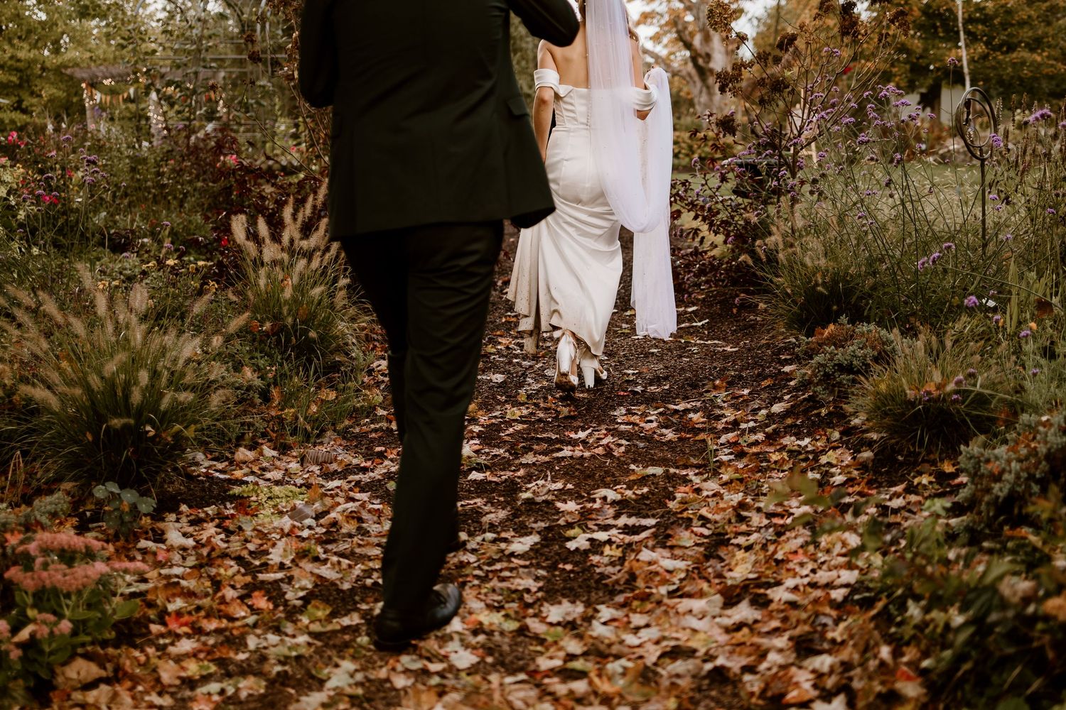Wedding couple walking on leaf-covered path through autumn garden.