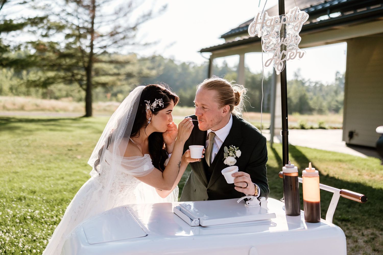 Bride and groom share a sweet moment tasting food at their outdoor wedding reception table on a sunny day.