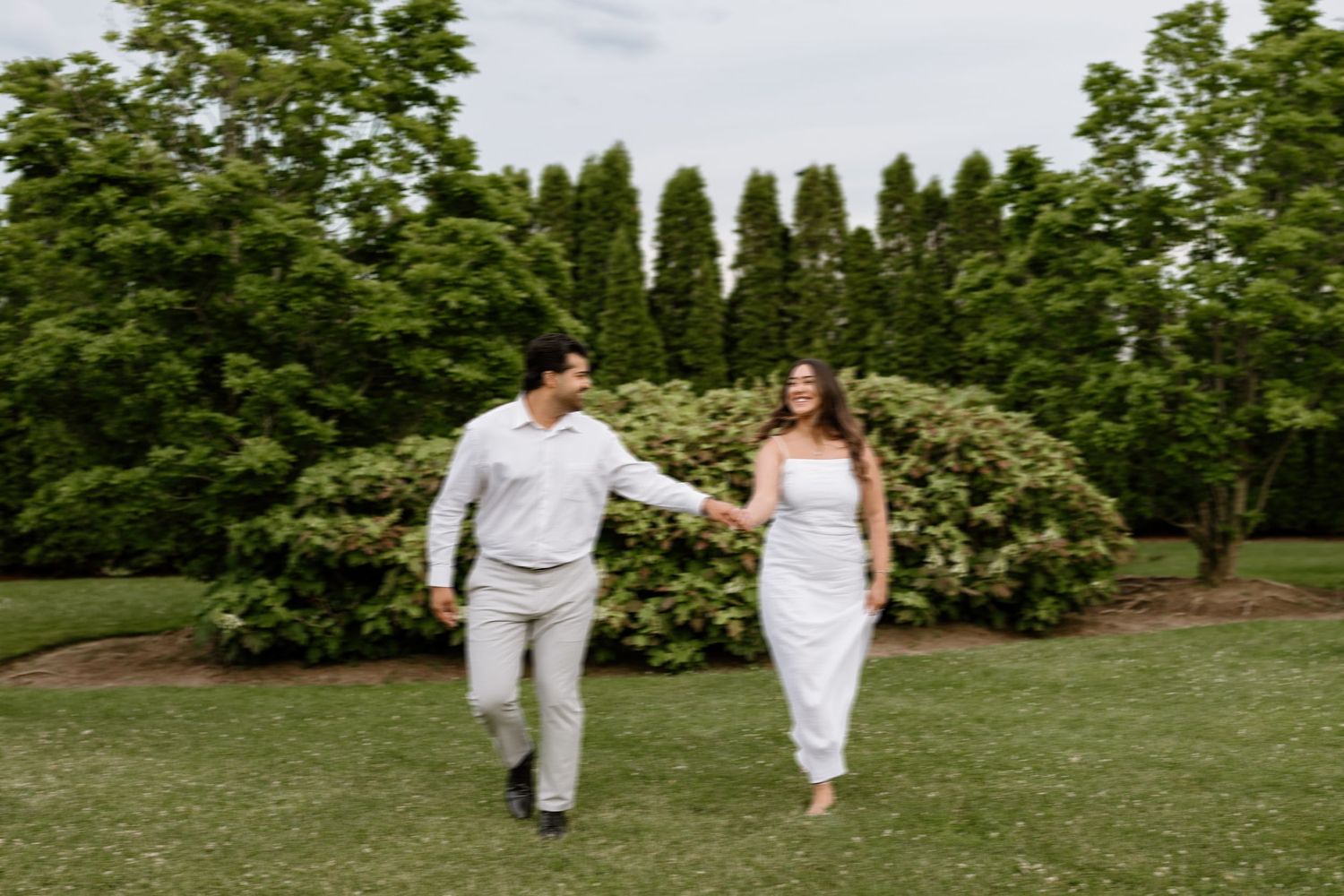A couple holds hands and walks across a lush green lawn surrounded by tall trees and blooming shrubs.