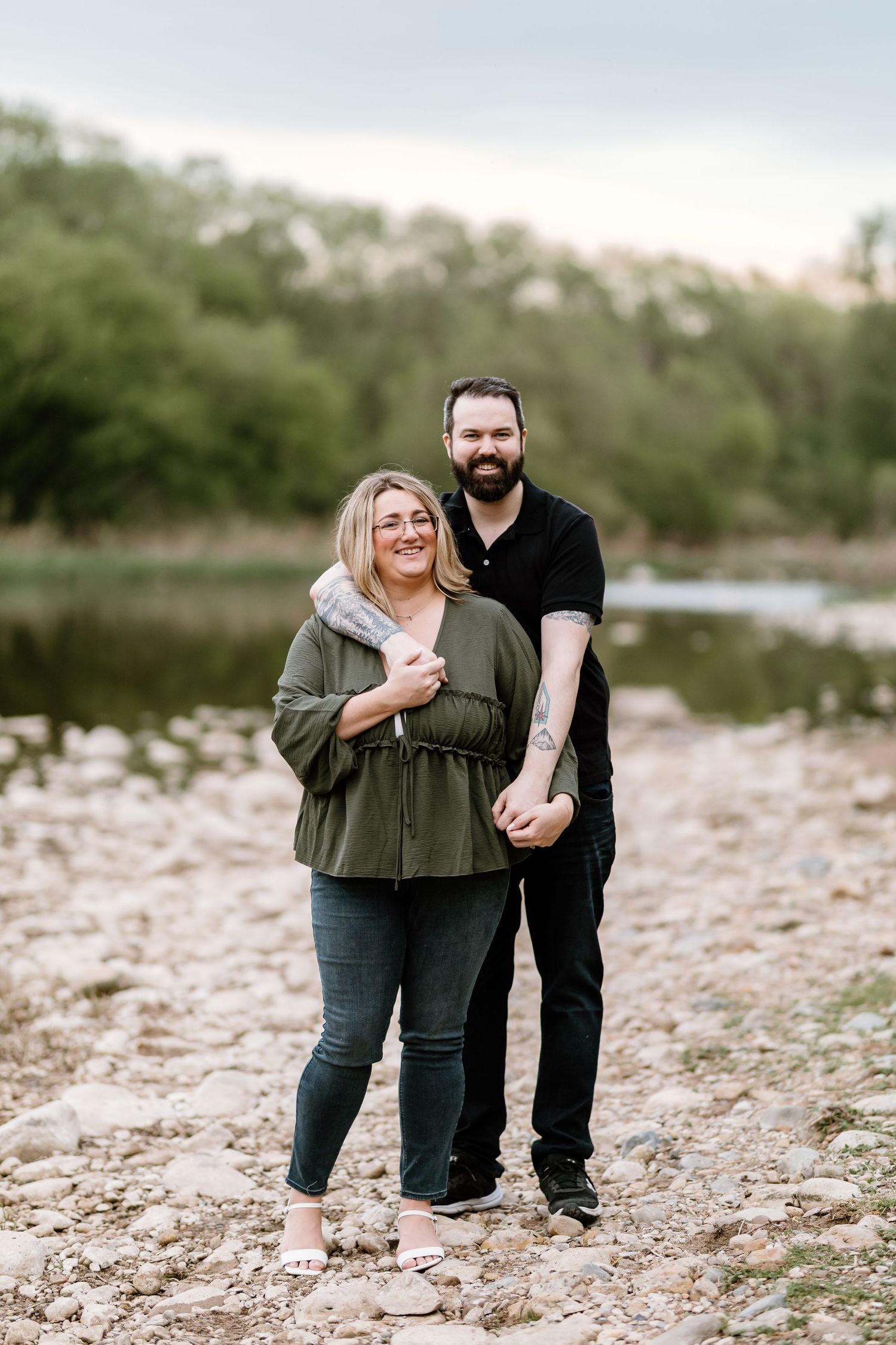 A smiling couple poses on rocky ground near a river, woman in green top and man in black shirt with a beard.