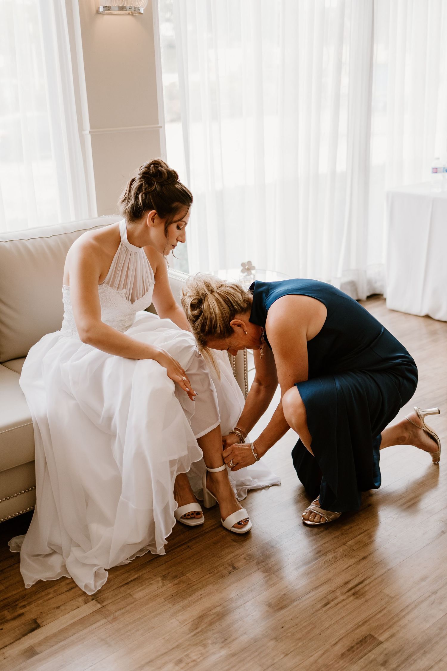 A person in a white dress getting help with shoes while seated on a beige couch.