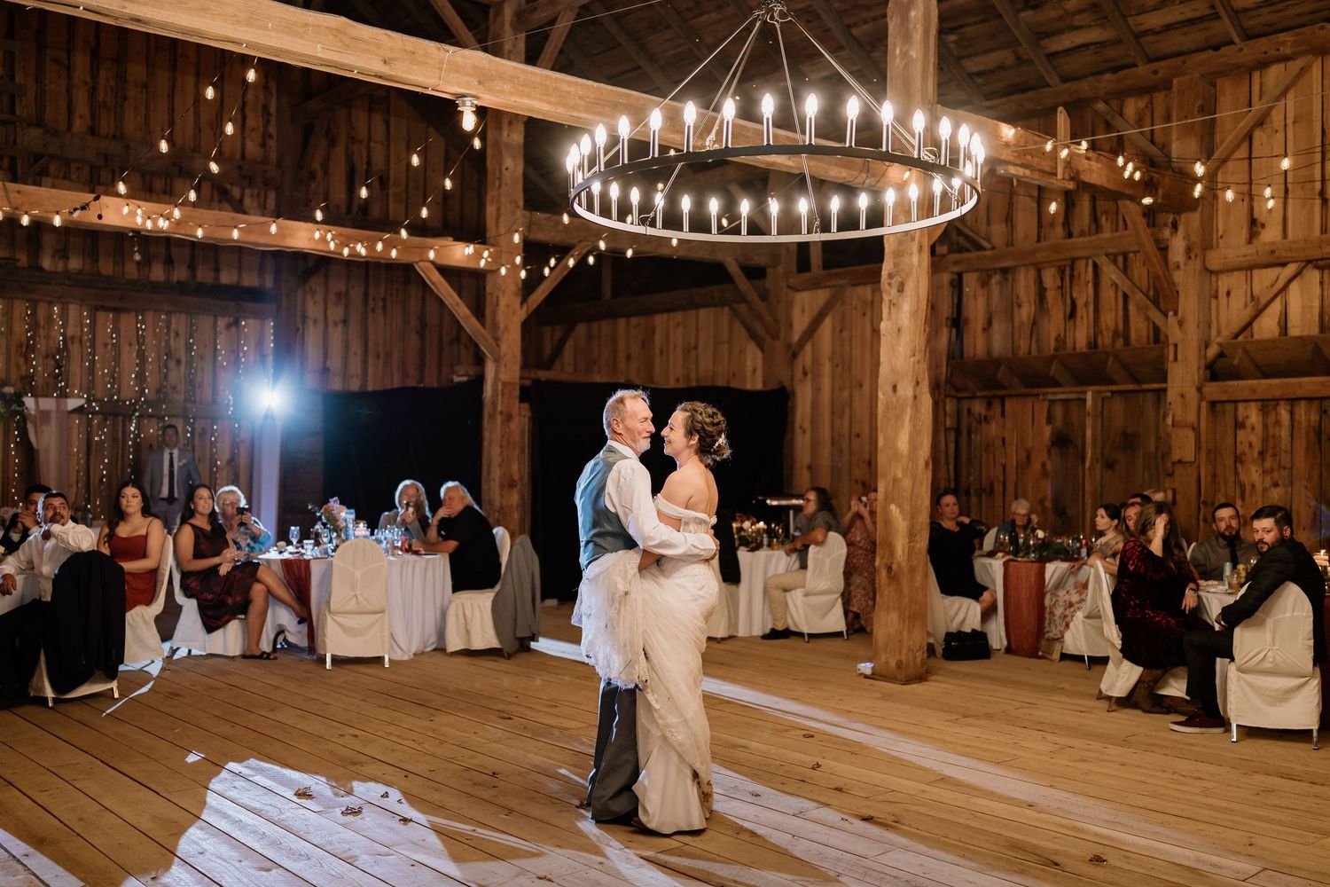 Couples dancing during a wedding celebration in a barn with wooden beams and circular chandelier overhead.