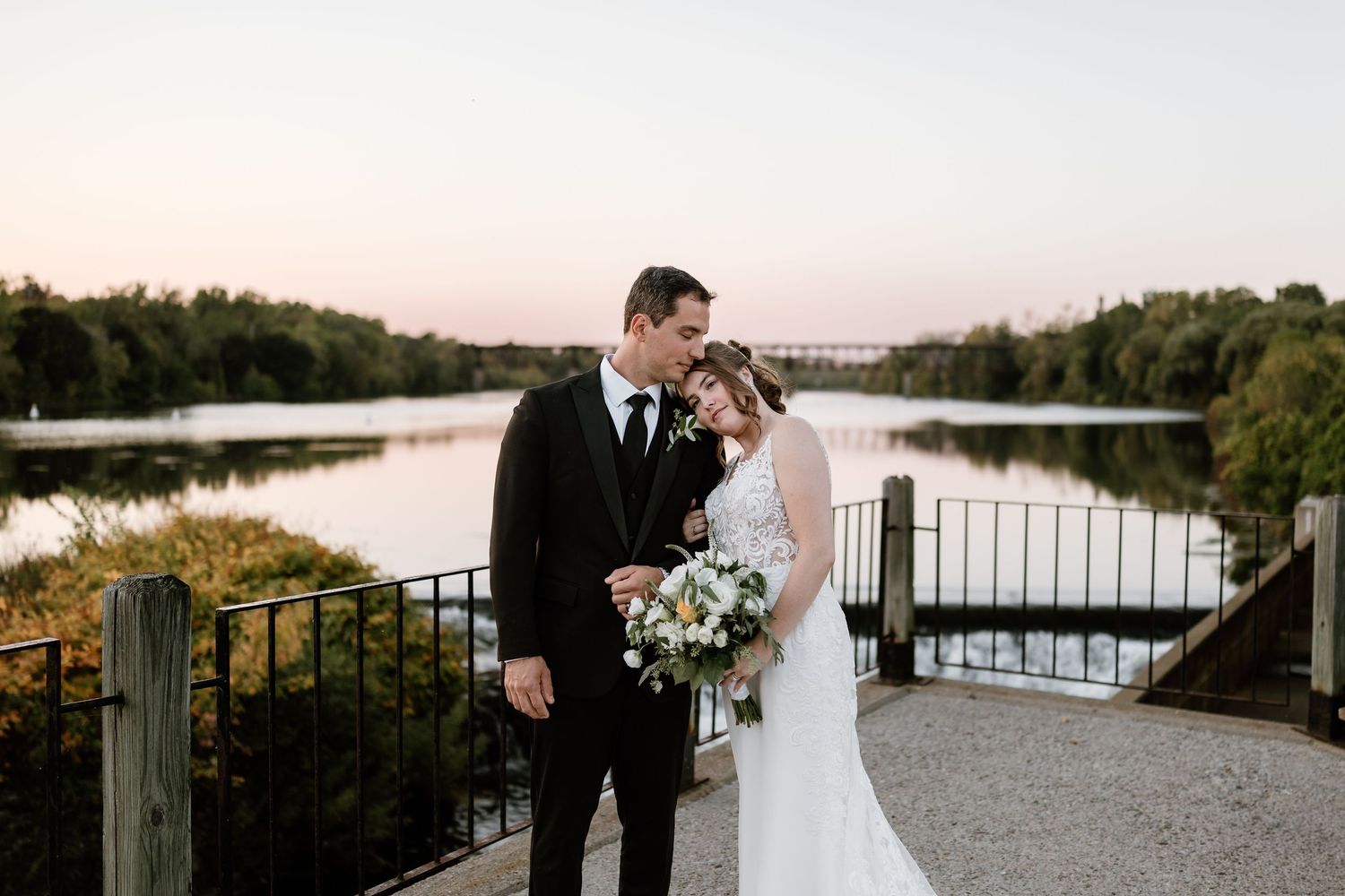 Wedding couple poses against sunset at lakeside pavilion with dramatic long train dress.