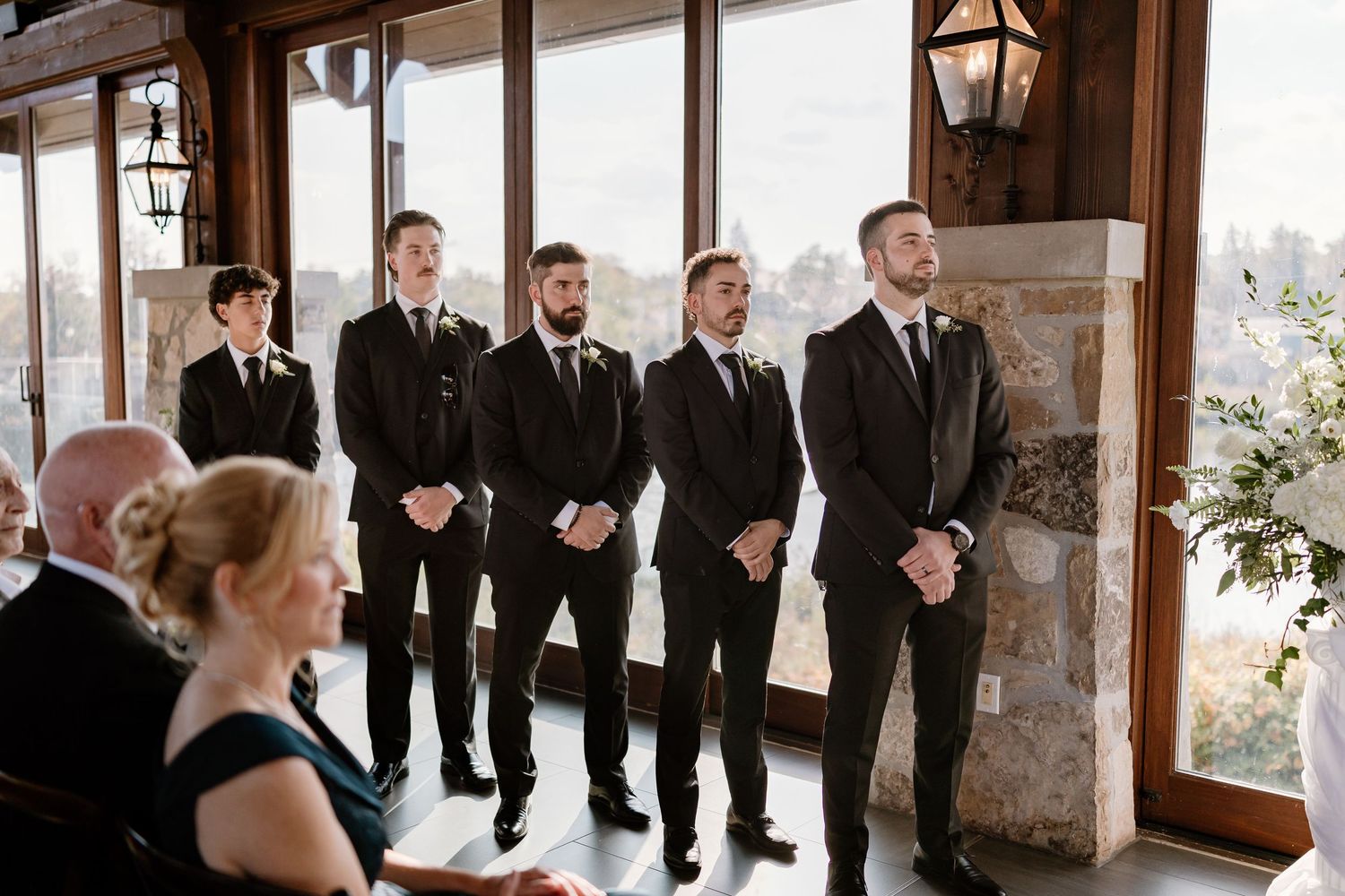 Groomsmen in black suits lined up during wedding ceremony by large windows.