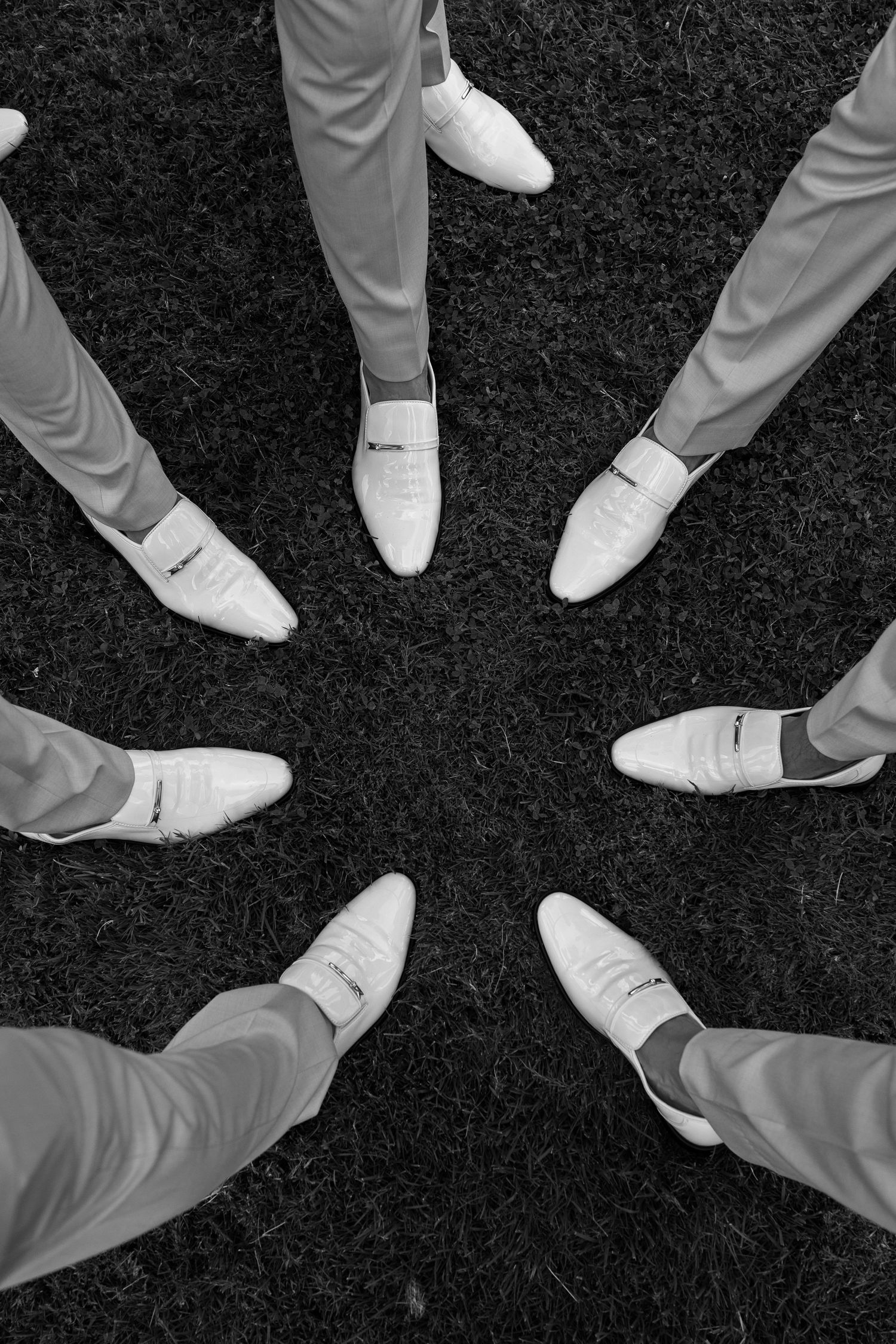 Black and white aerial view of groomsmen in gray suits forming a circle with white dress shoes on grass.