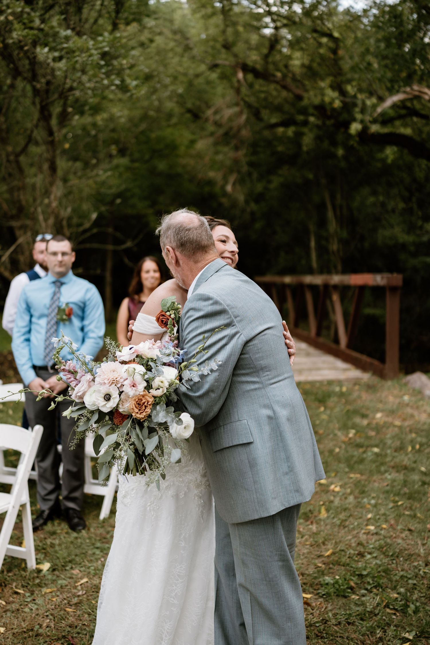 Intimate outdoor wedding ceremony moment with guests watching couple embrace near white chairs and floral decor.