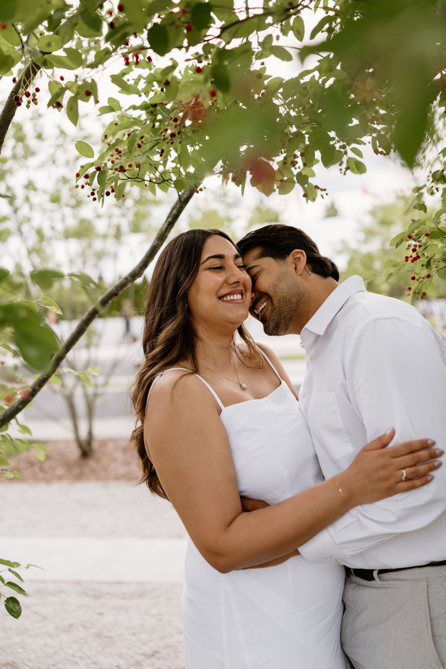 Couple embracing under berry tree branches, woman in white dress smiling as man whispers in her ear outdoors.