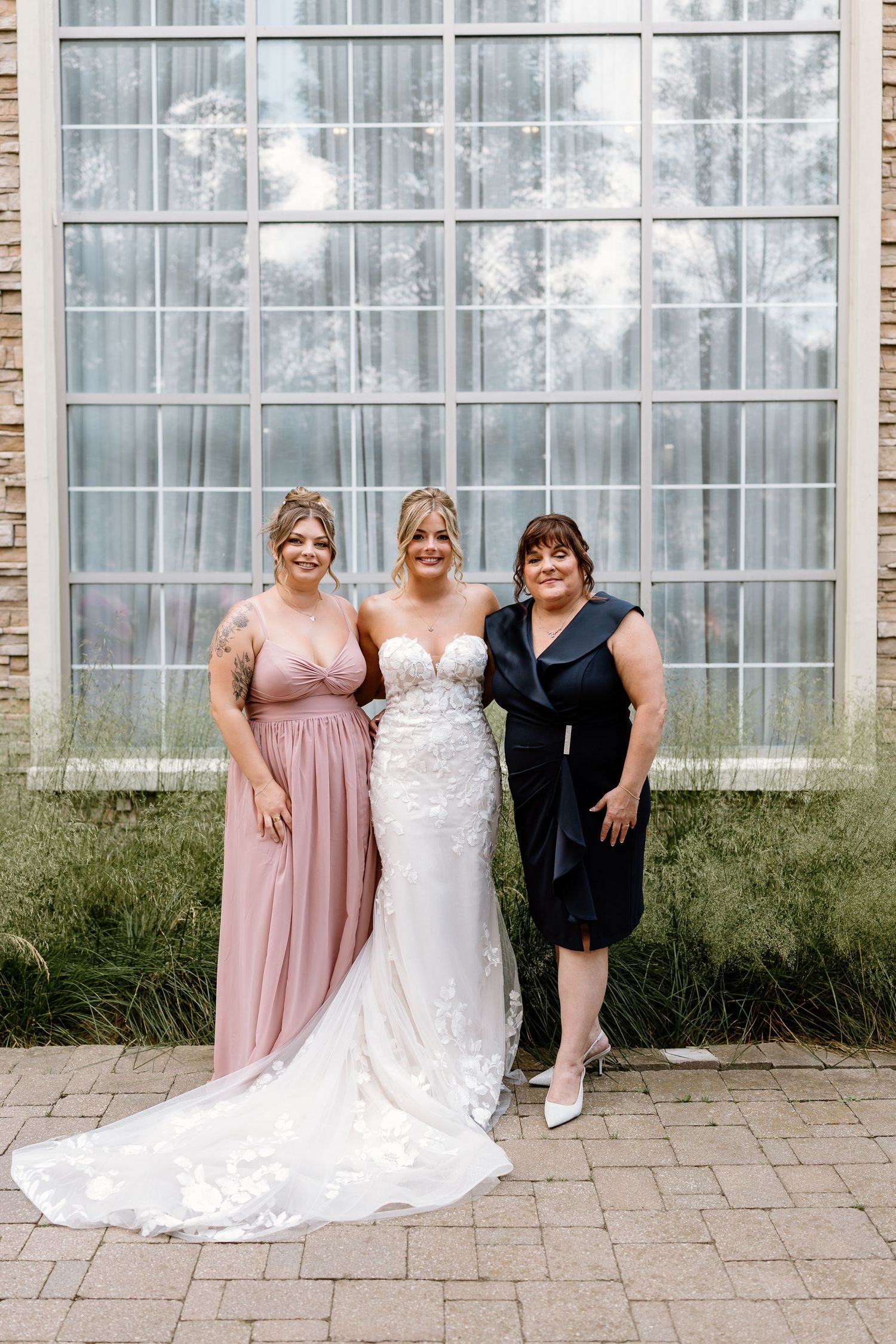 Bride in white lace gown poses with bridesmaid in pink dress and guest in navy blue outside a modern building.