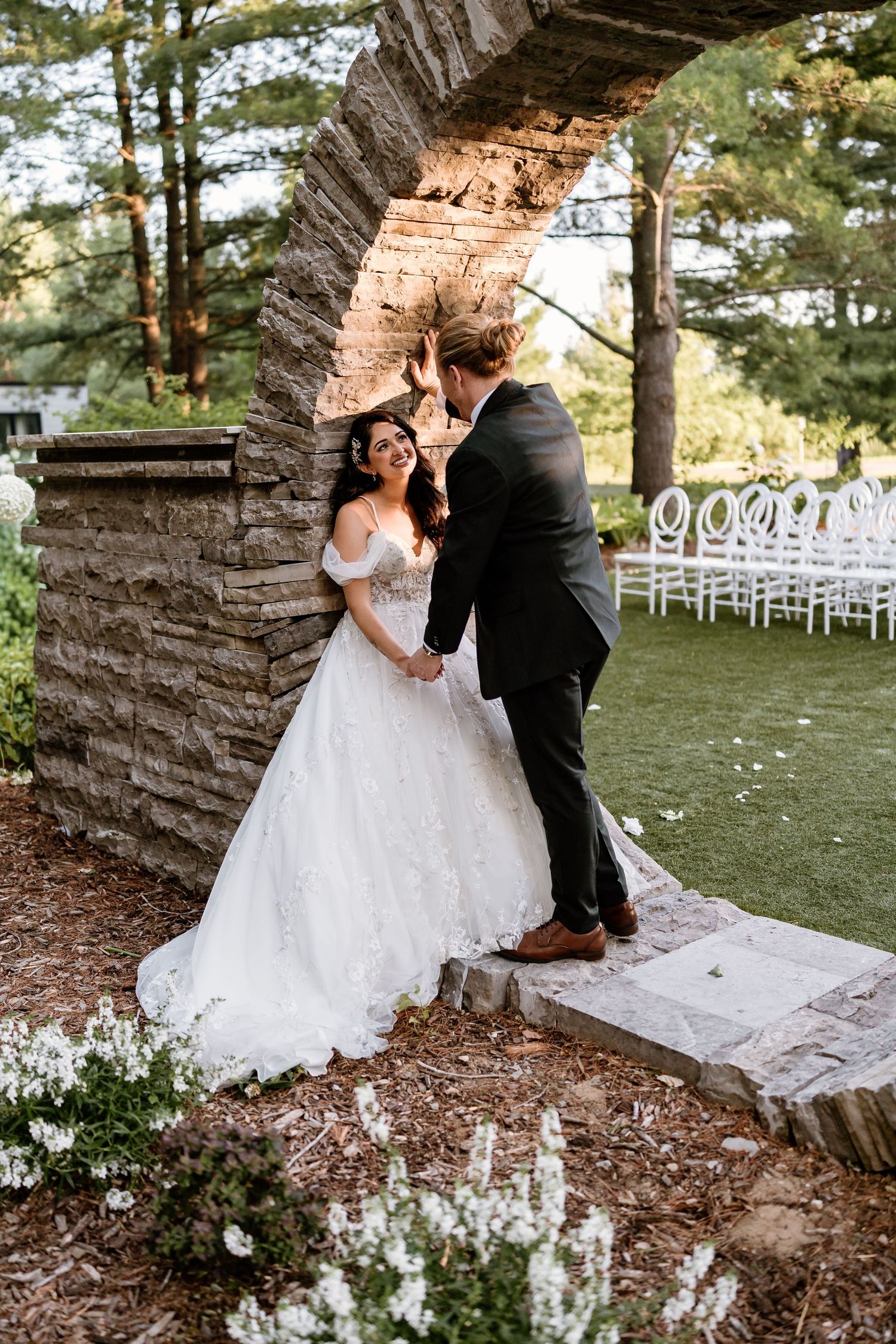 Bride and groom share a romantic moment under a stone arch at an outdoor wedding venue surrounded by lush greenery.