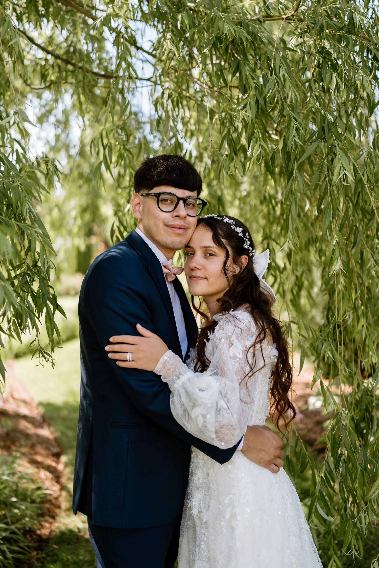 A bride and groom embrace under a weeping willow tree, she in a lace gown and he in a navy suit with pink bow tie.