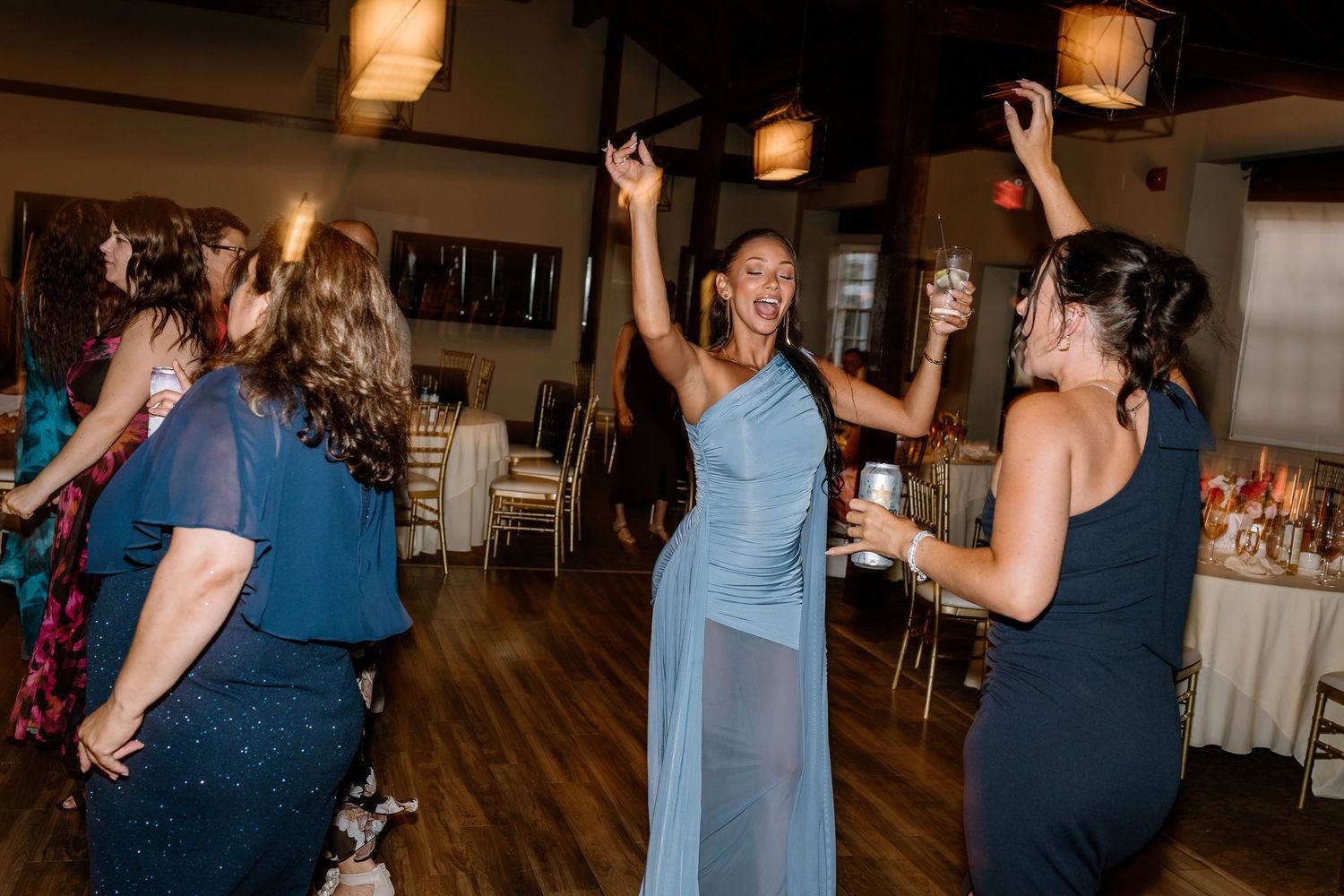 Guests in elegant dresses dance joyfully at an indoor wedding reception celebration.