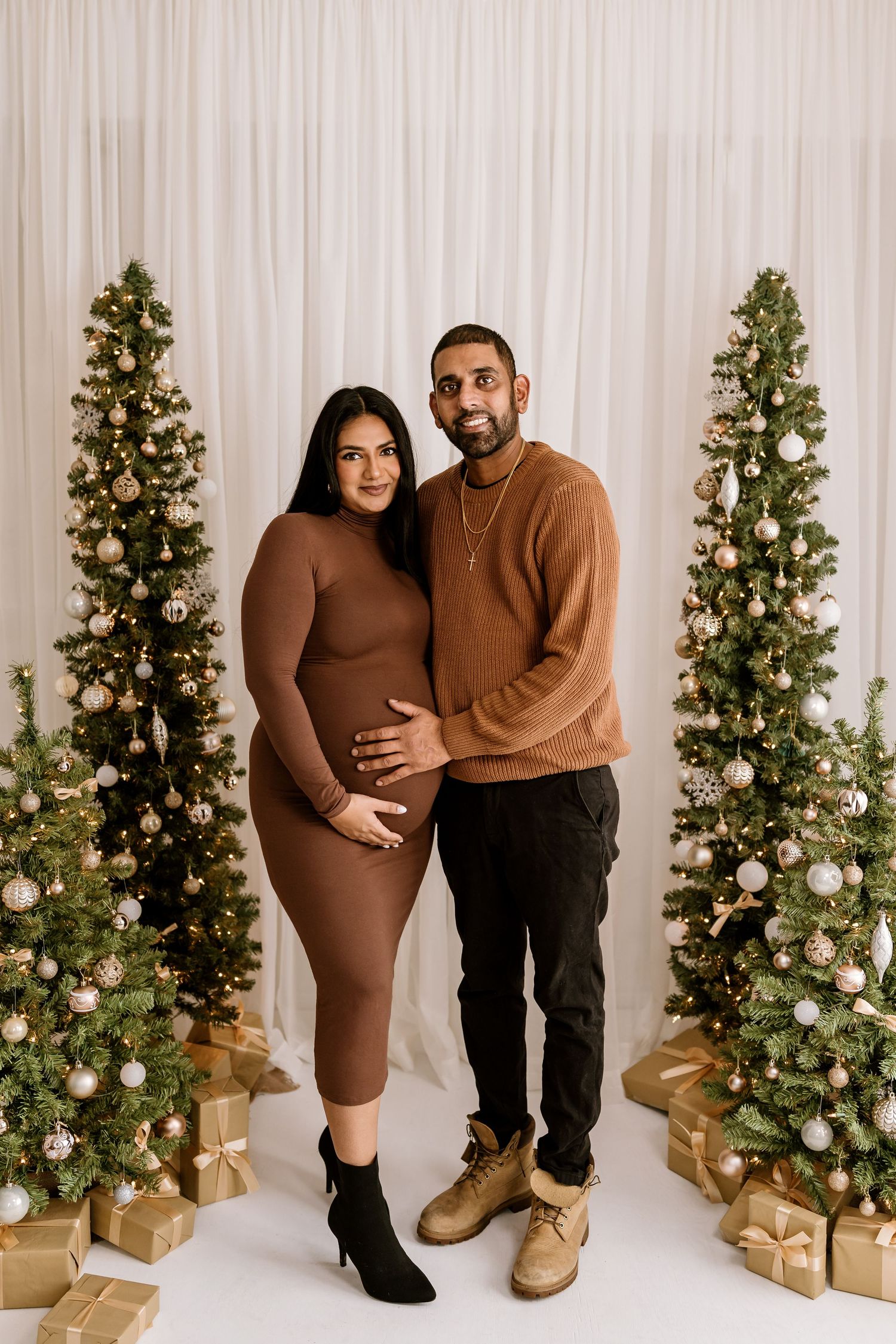 Holiday maternity photoshoot with decorated Christmas trees and wrapped presents in background.