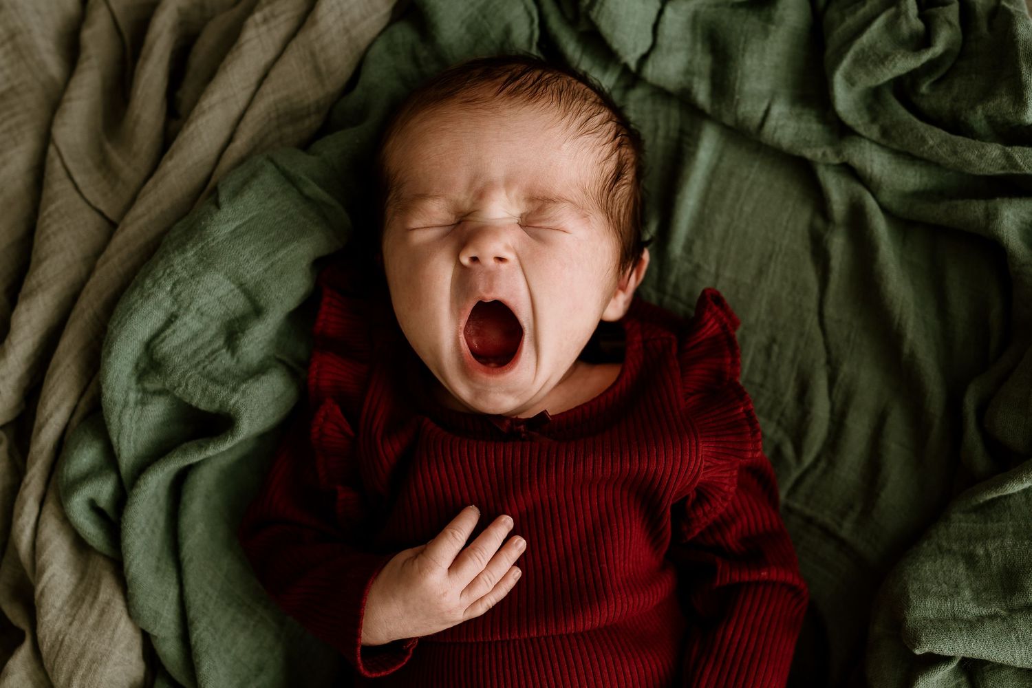 Newborn baby wearing red clothing yawns while lying on soft green blanket in artistic photo series.