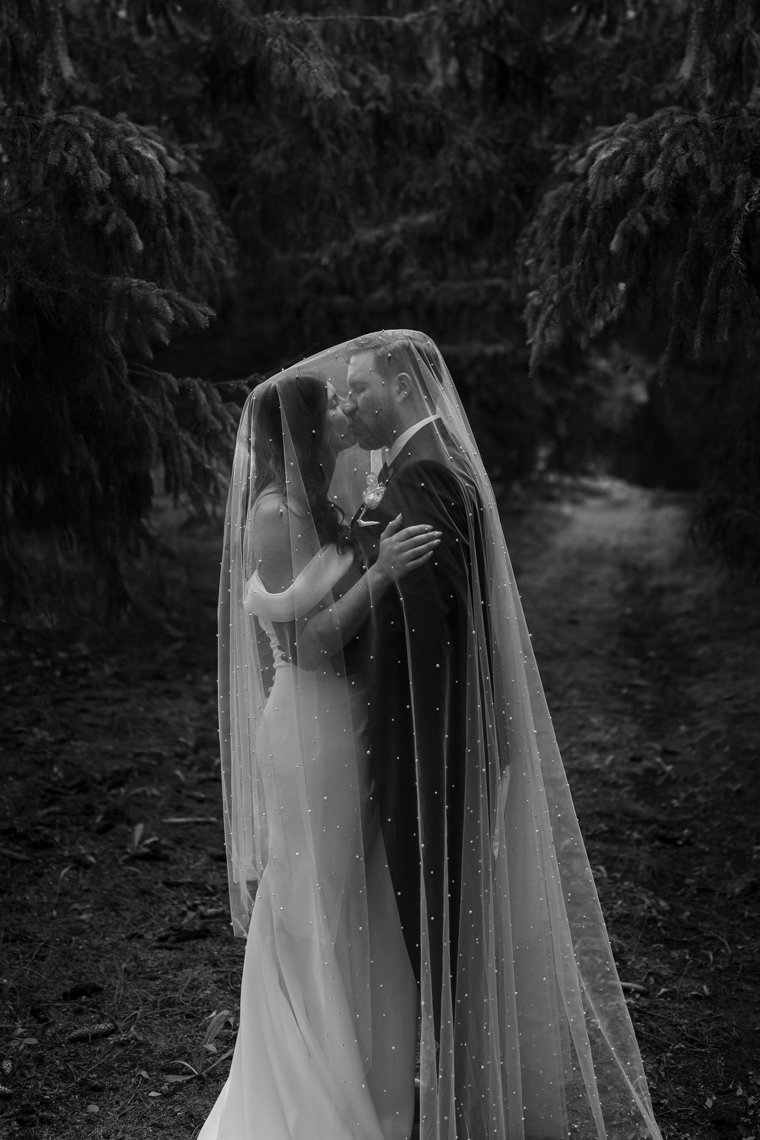 Romantic wedding couple embraces under flowing white veils in a wooded forest setting.