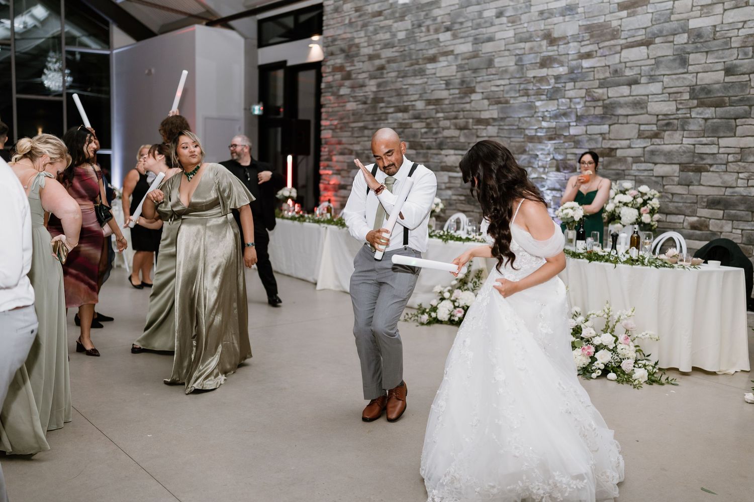 Bride and groom dancing joyfully at wedding reception with guests in sage green bridesmaid dresses celebrating around them.