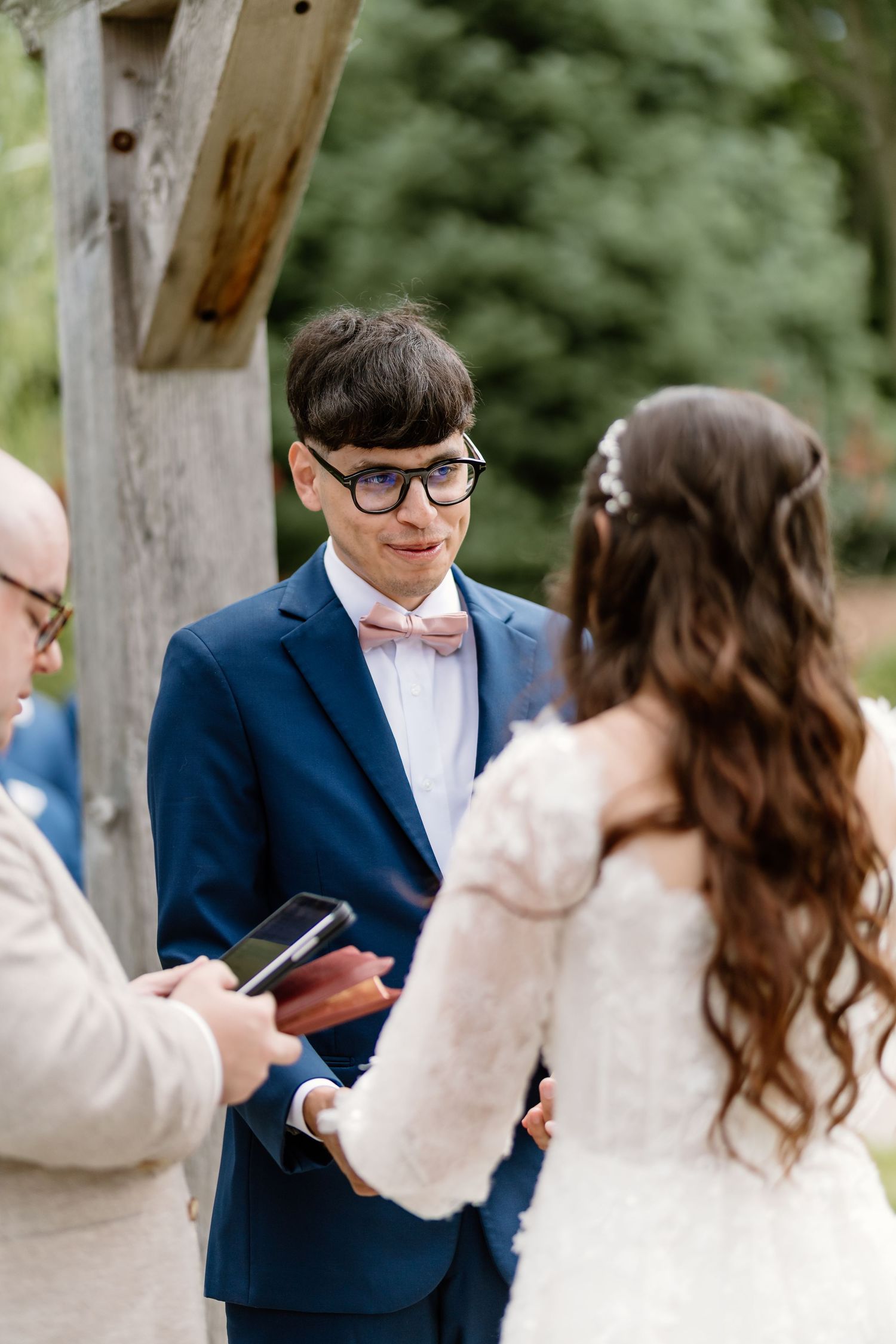 Groom in blue suit and pink bow tie exchanges vows with bride during outdoor wedding ceremony.