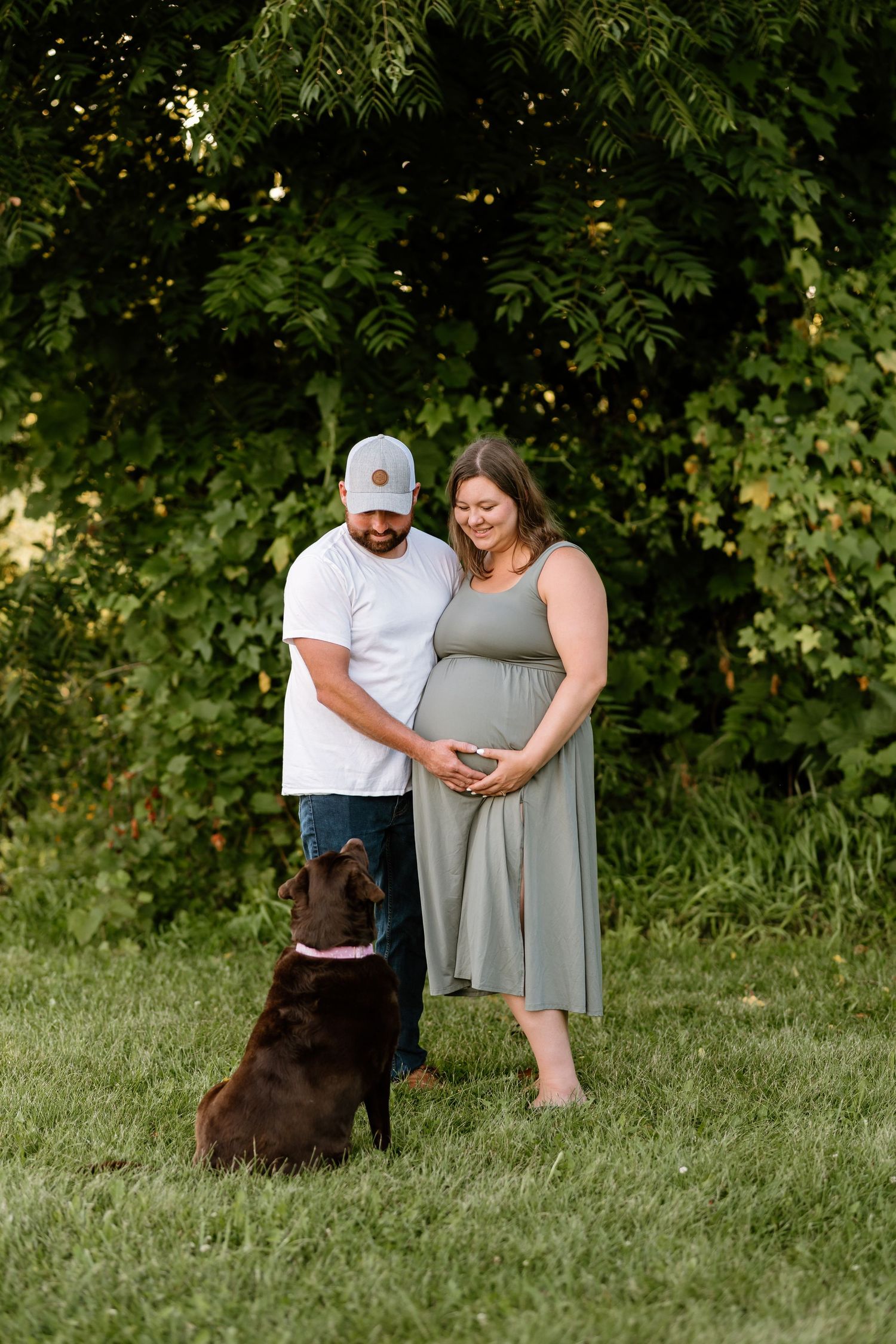 Pregnant couple in green field with chocolate lab dog, woman in sage dress holding baby bump, man in white cap and tee.