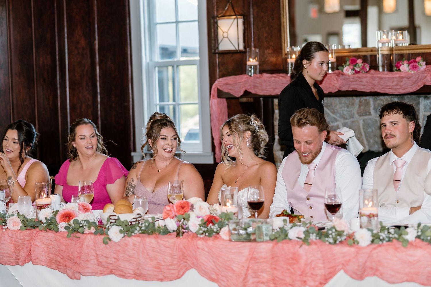 Wedding party seated at a decorated head table with pink linens, floral arrangements, and candles at a reception.