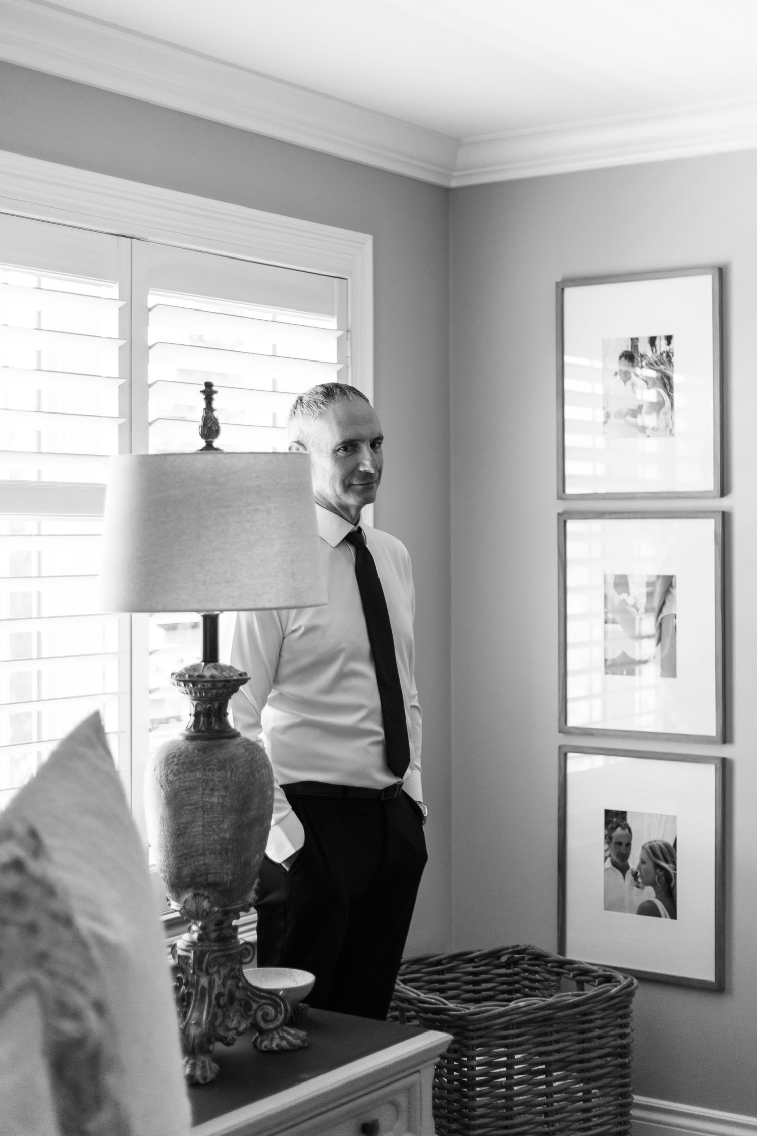 Black and white portrait of person in formal attire standing by window with table lamp.