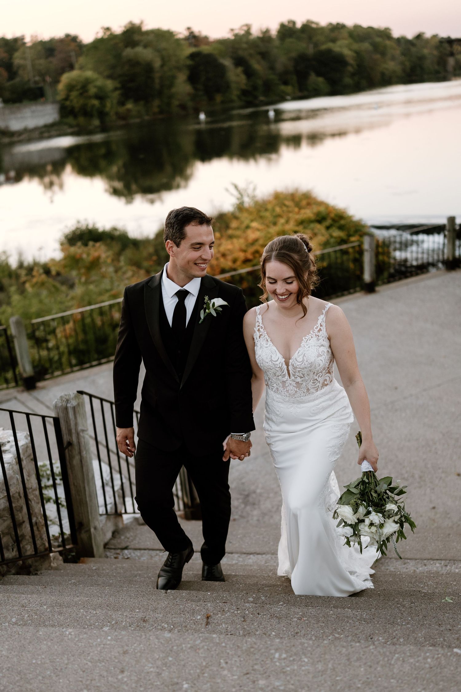 Newlyweds share joyful moment while walking along waterfront path at sunset.