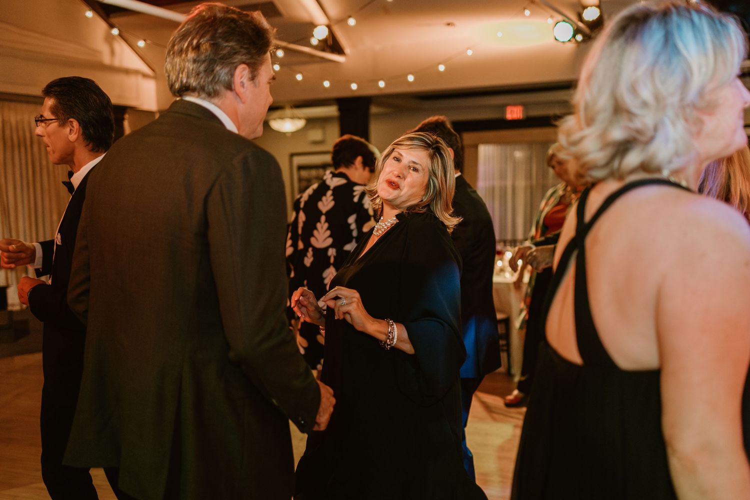 Wedding guests mingling and dancing at an indoor reception with string lights overhead.