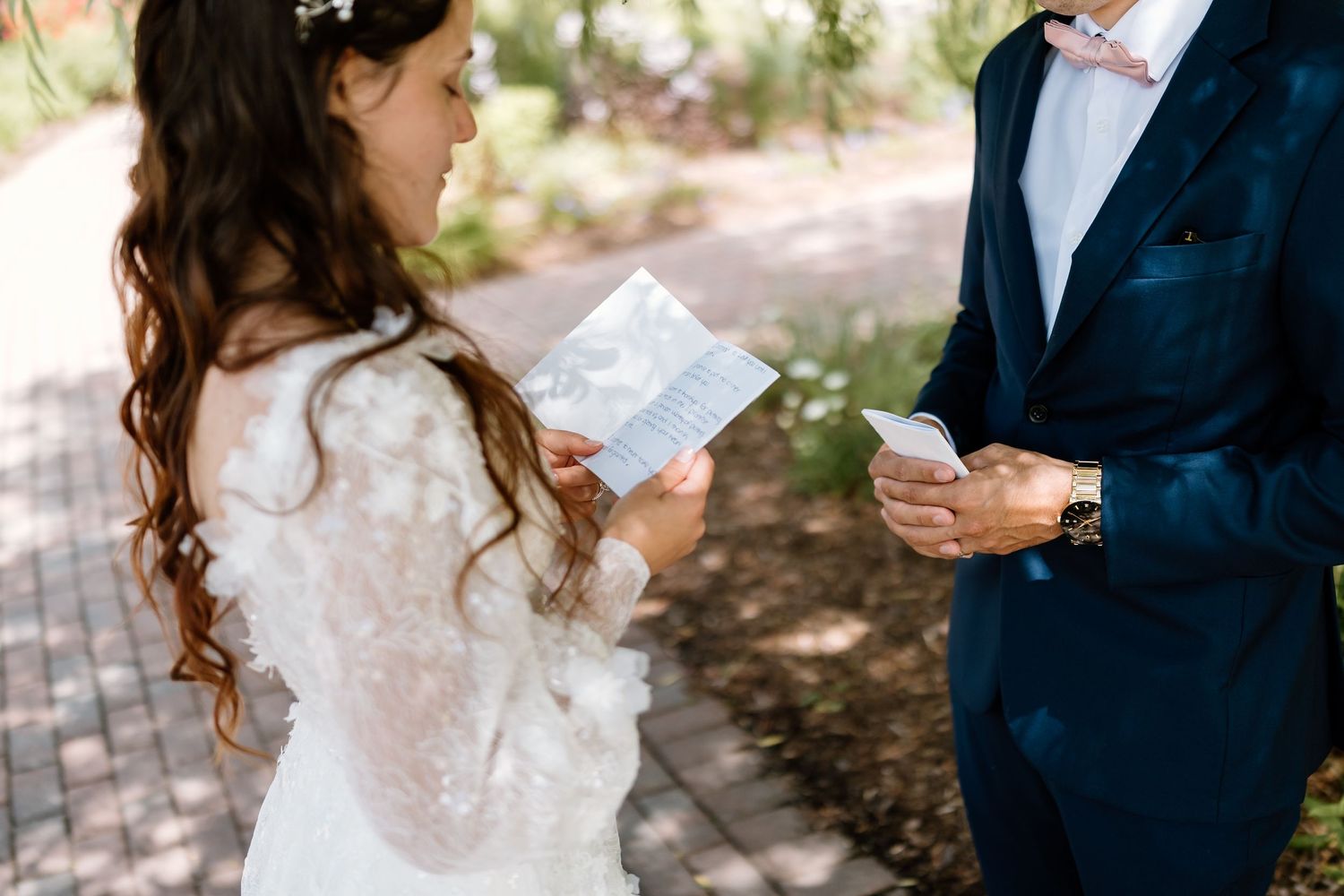 Bride in lace dress reads vows from a card while groom in navy suit listens during outdoor wedding ceremony.