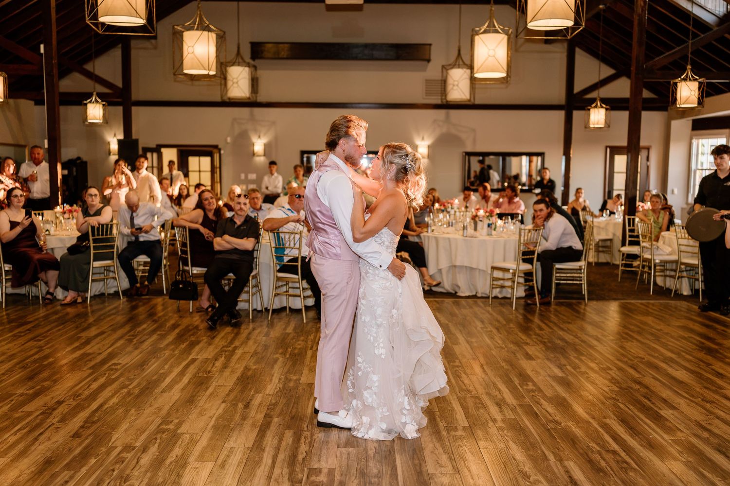 Bride and groom share first dance in elegant rustic wedding reception hall with warm pendant lighting and seated guests.