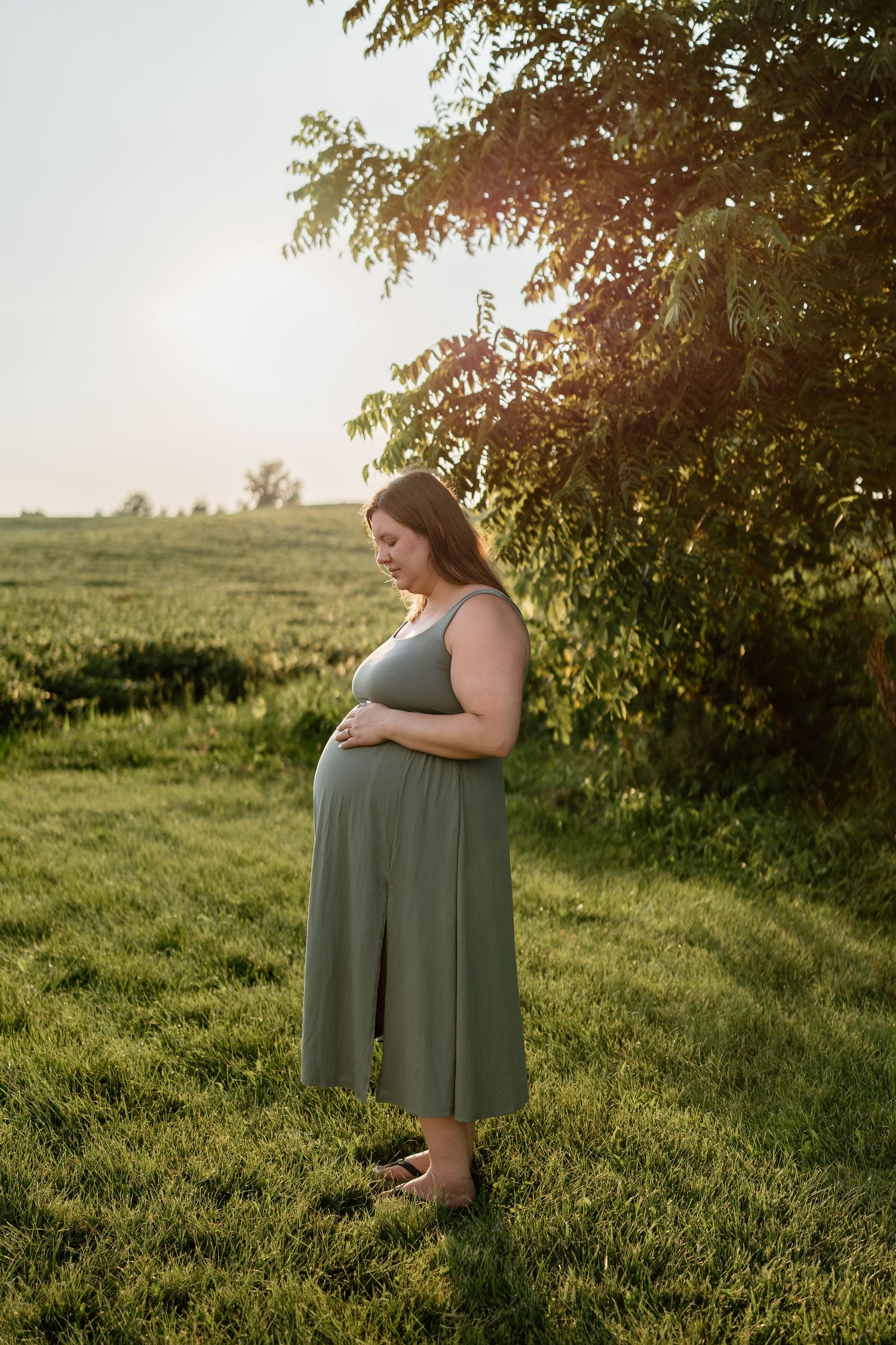 Pregnant woman in olive green midi dress standing in sunlit green field, cradling her baby bump near a tree at golden hour.