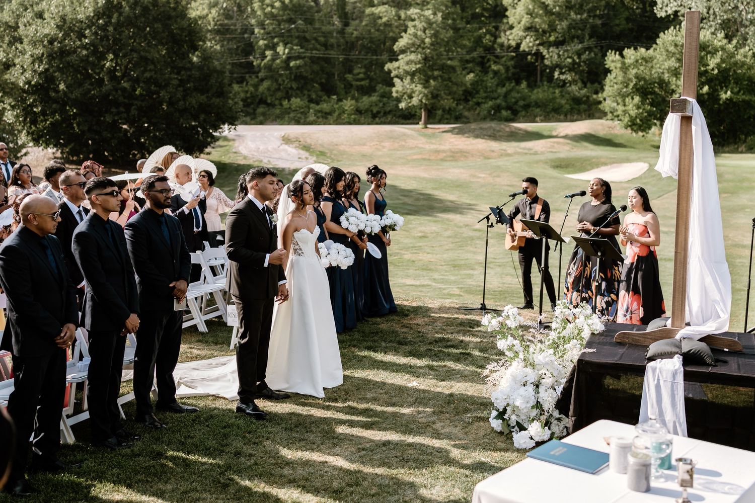 Outdoor wedding ceremony with guests seated in white chairs on a sunny day.