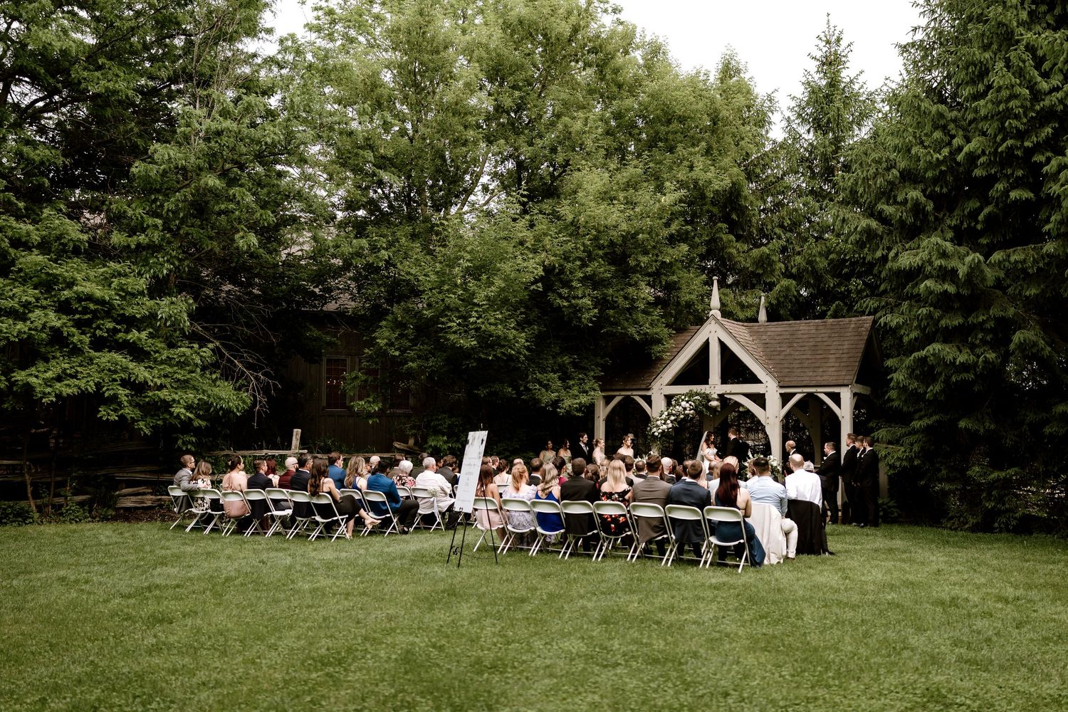 Outdoor wedding ceremony setup with wooden gazebo and rows of white chairs on lush green lawn.