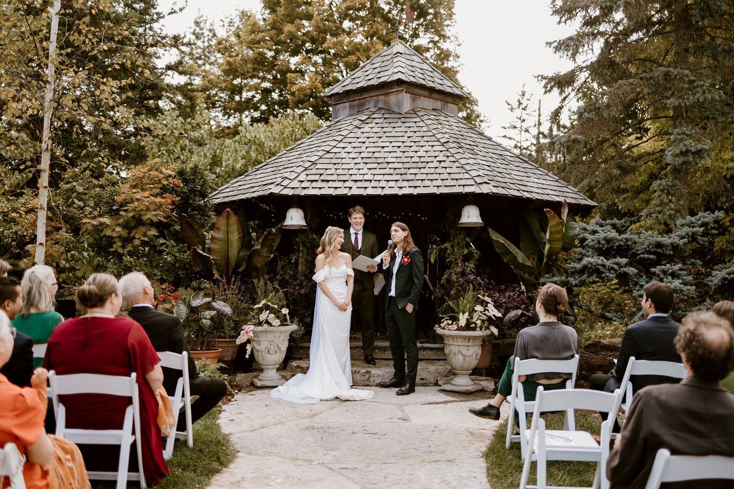 An outdoor wedding ceremony takes place under a wooden gazebo surrounded by trees and white chairs.