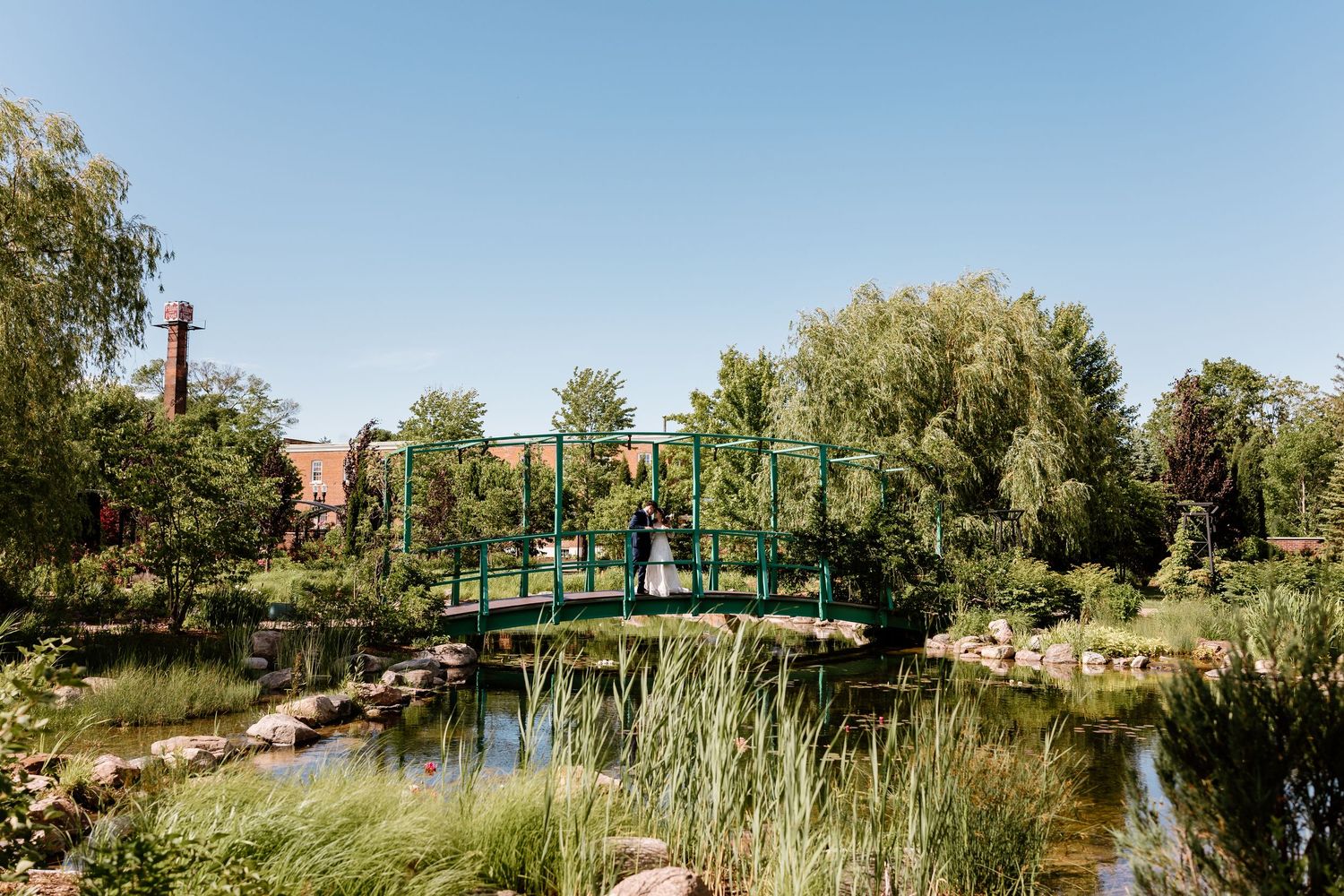 Couple standing on a green bridge over a tranquil pond surrounded by lush greenery under a clear blue sky.
