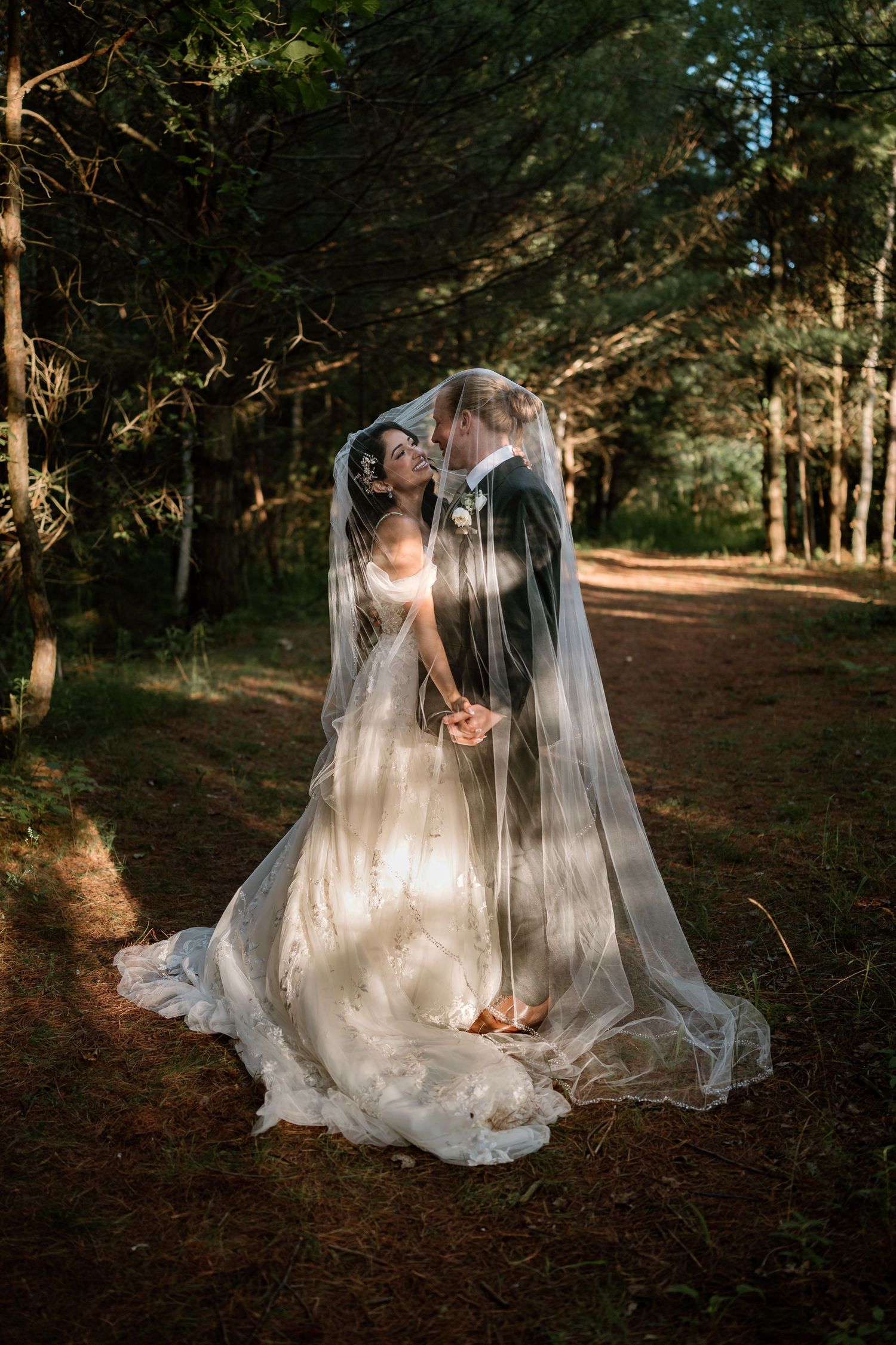 A bride and groom share a romantic kiss under a flowing veil in a sunlit forest setting.