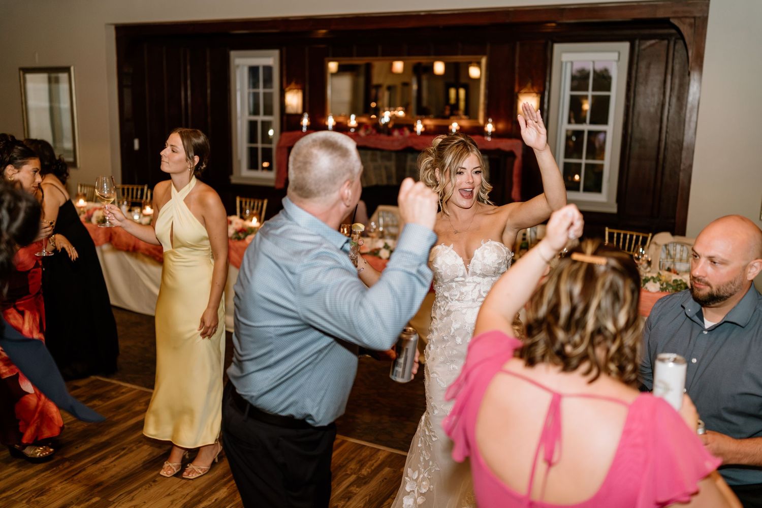 Bride in a lace gown dances energetically with guests at a lively wedding reception.