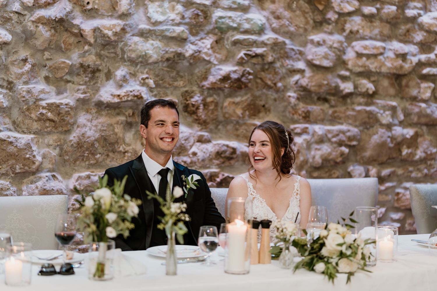 Wedding guests raise wine glasses in a toast at an elegant reception with stone walls and floral centerpieces.