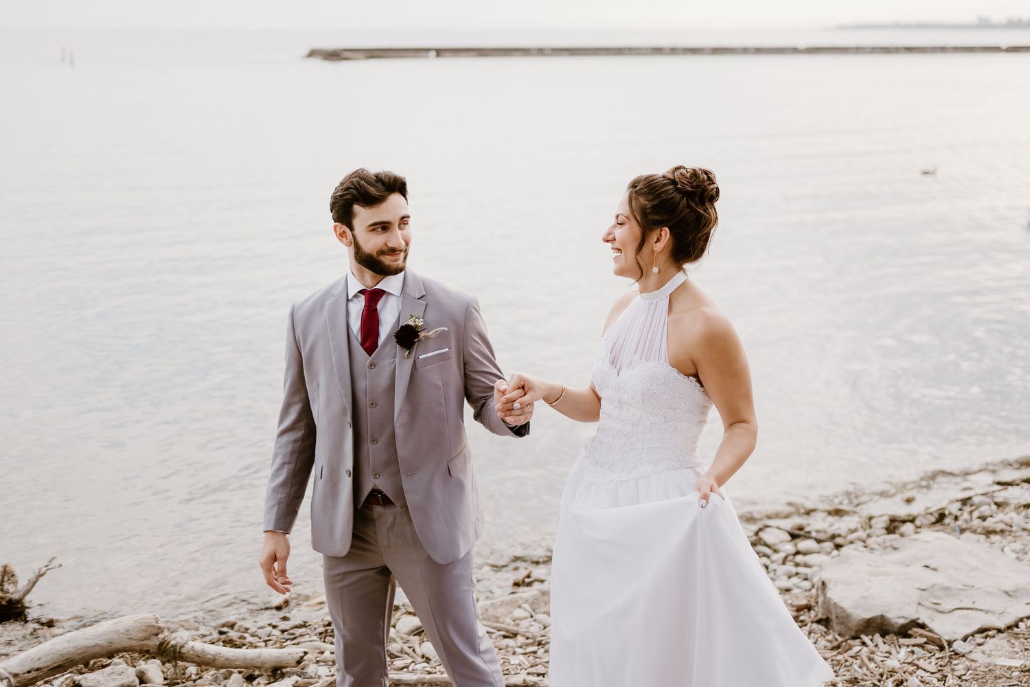 A joyful wedding couple shares a playful moment on a beach with water in the background.