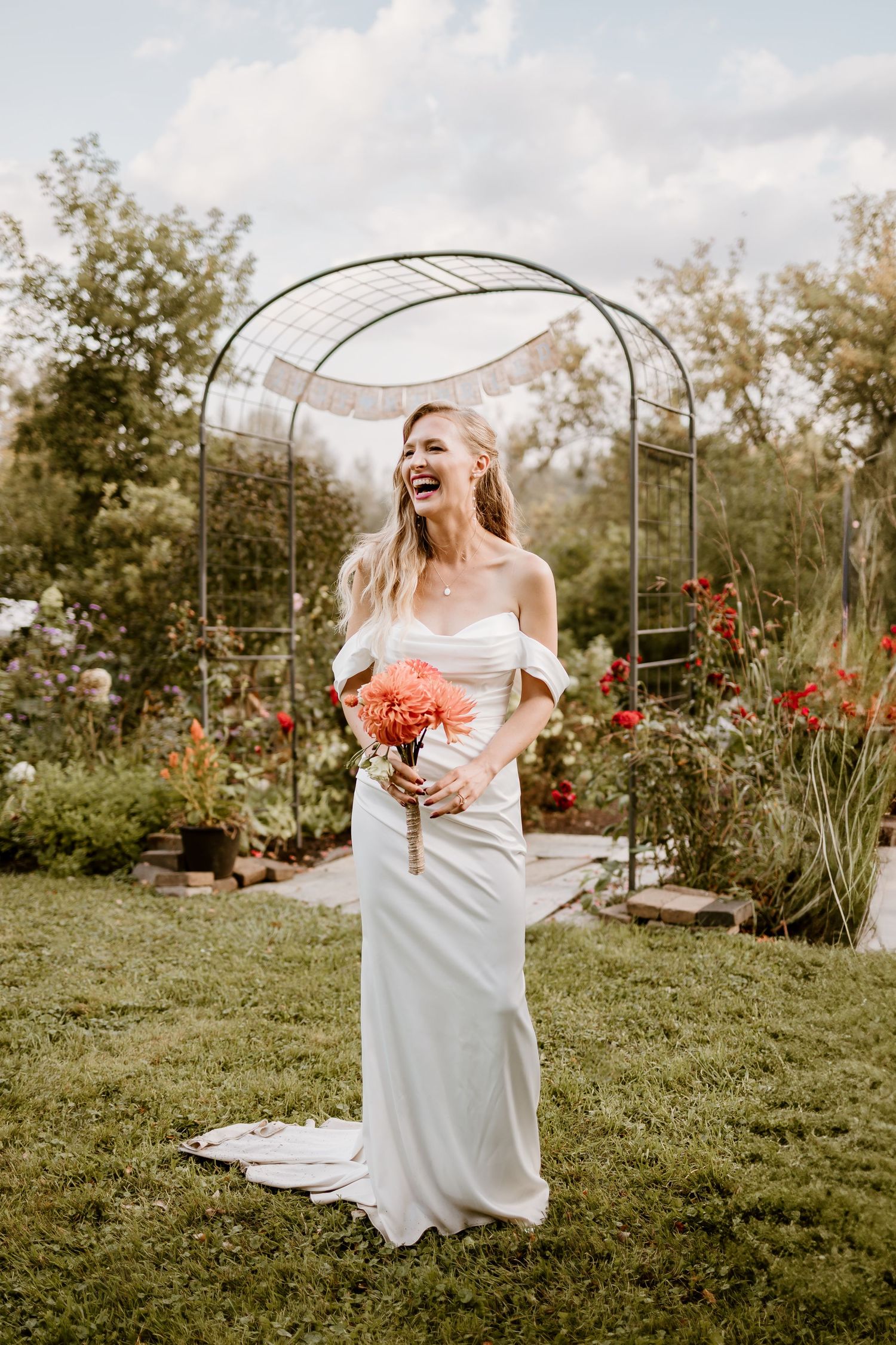 Wedding dress and coral bouquet under garden arch surrounded by blooming flowers.