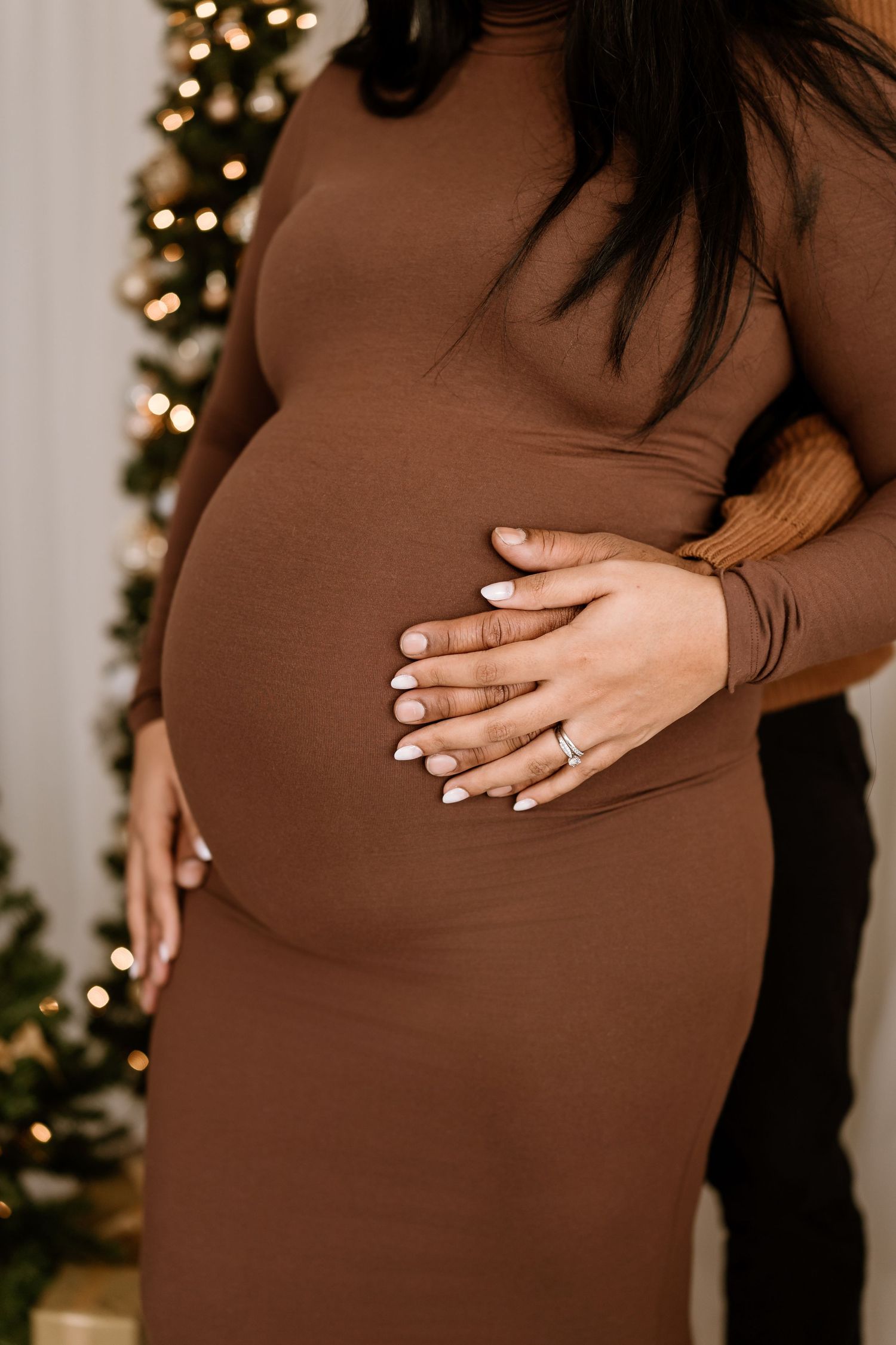 Close up of hands gently resting on a brown maternity dress against a festive Christmas backdrop.