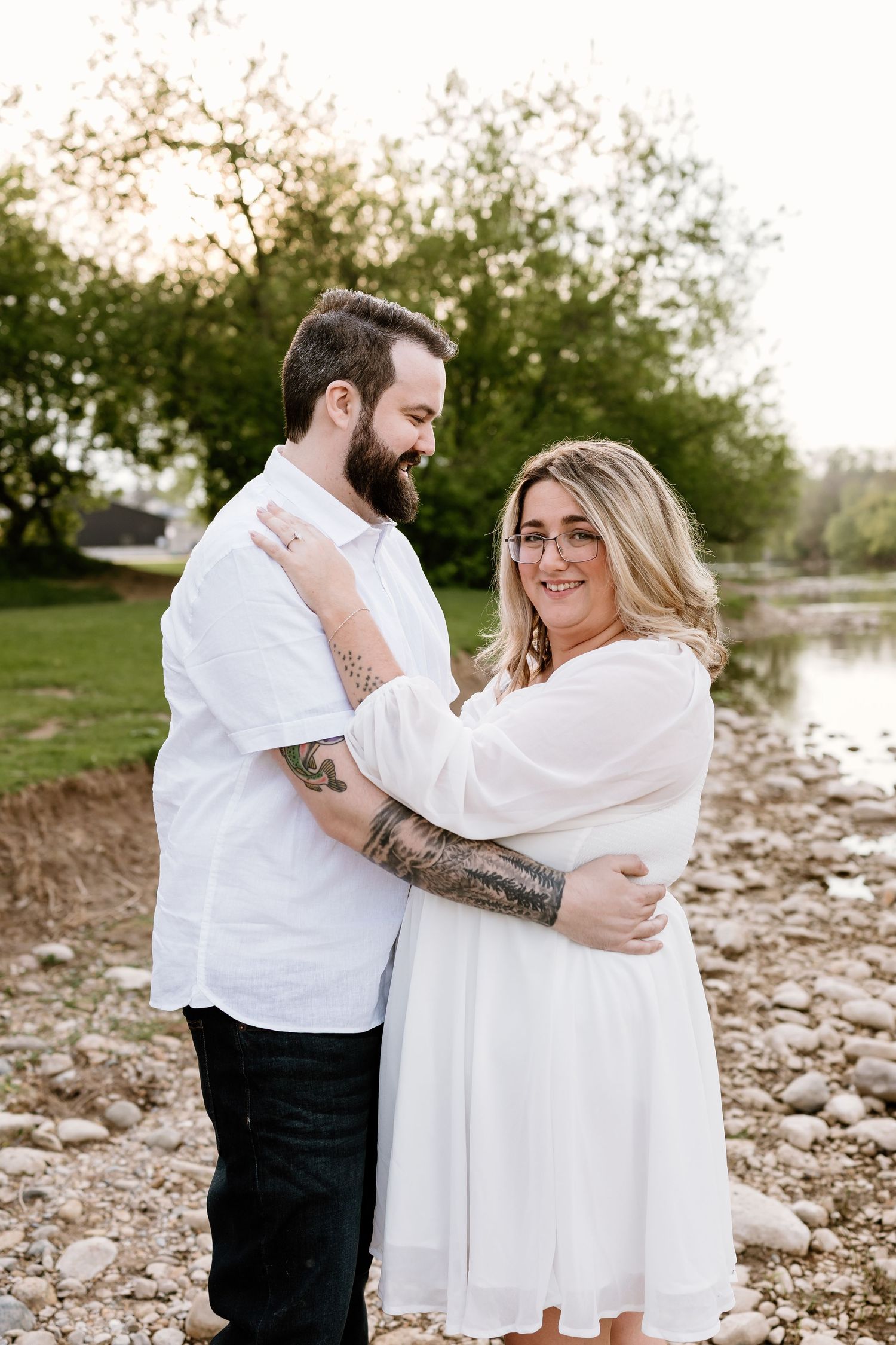 Couple embracing outdoors near rocky shoreline, woman in white dress smiling at camera, man with tattoos in white shirt.