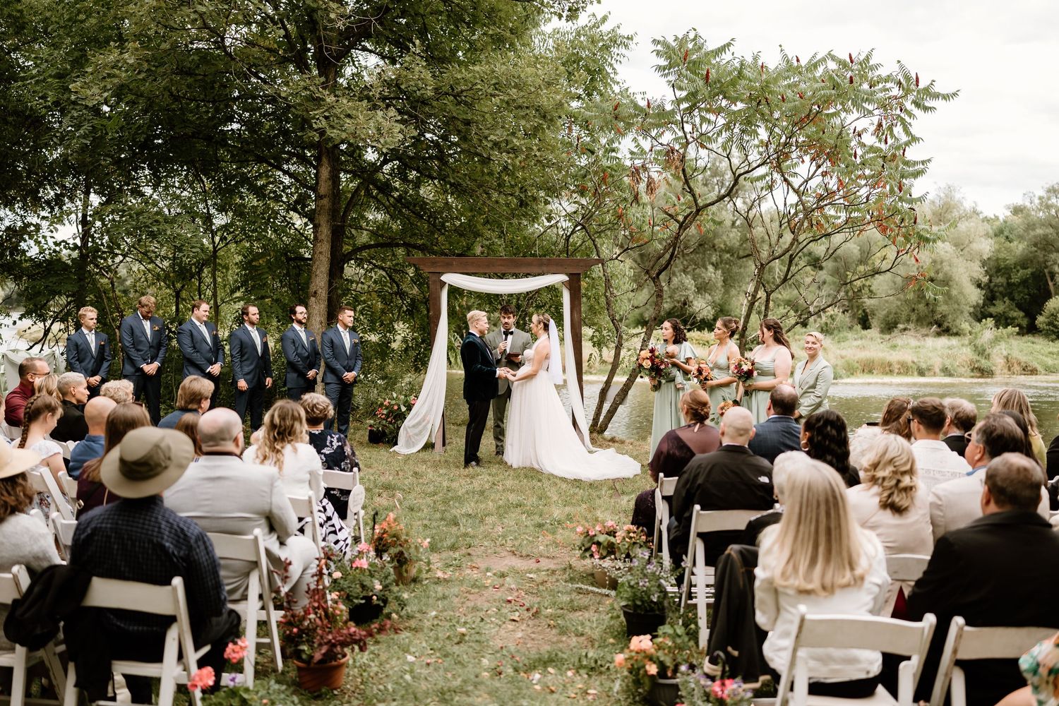 Wedding guests gather outdoors for a celebration ceremony under trees on a sunny day.