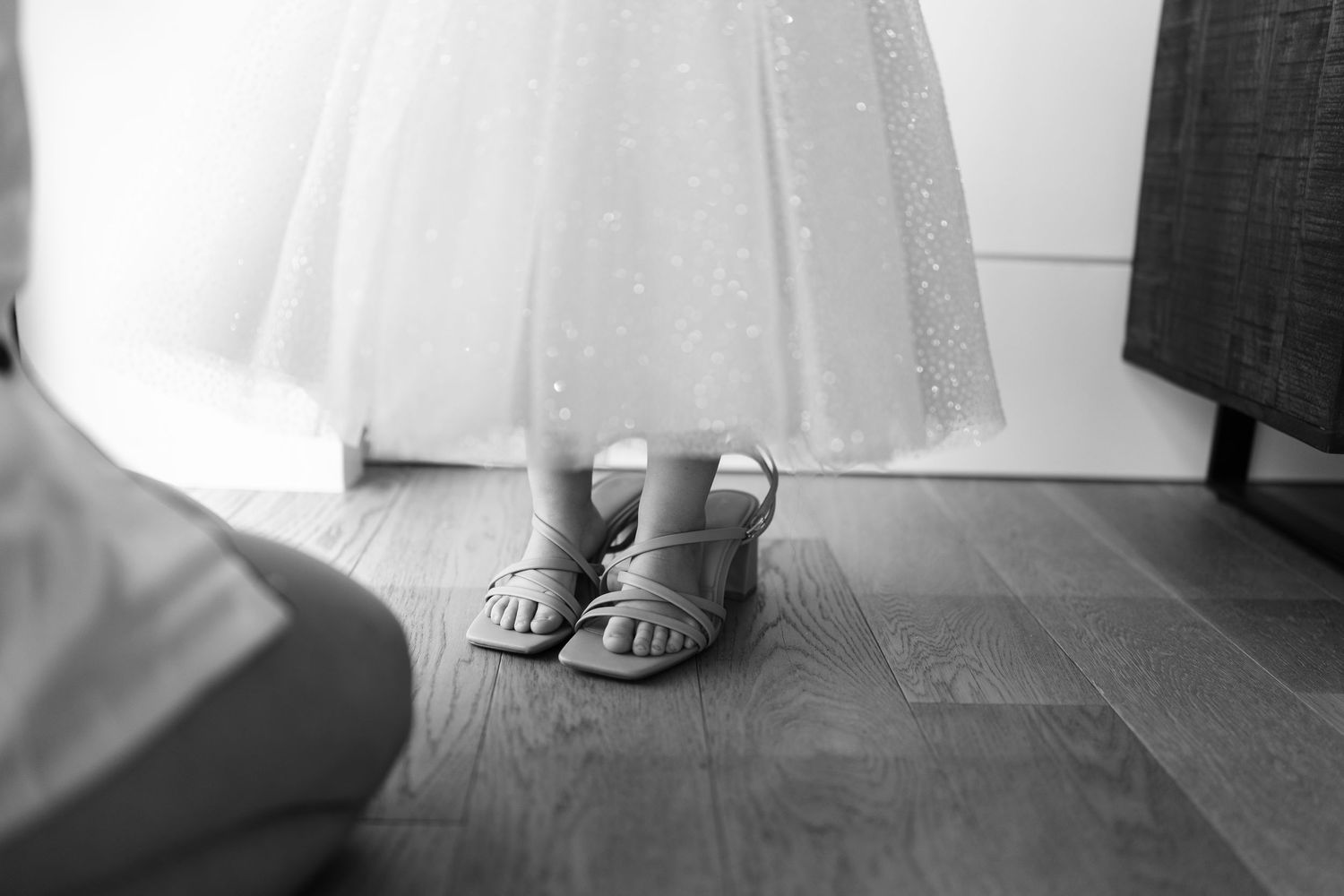 Close-up of small silver sandals on hardwood floor next to white dress hem.