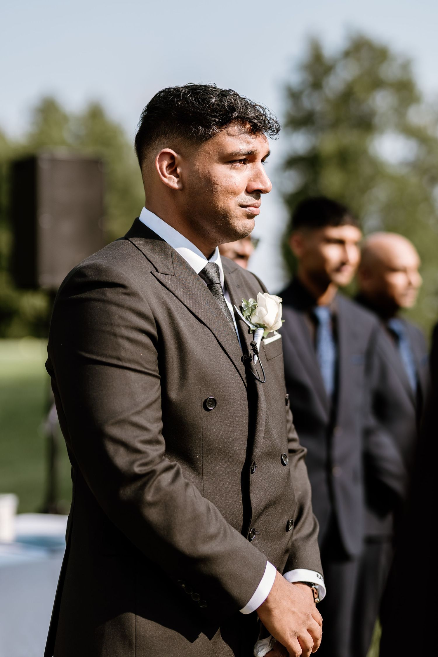A groom in a brown suit with boutonniere stands outdoors during a wedding ceremony.