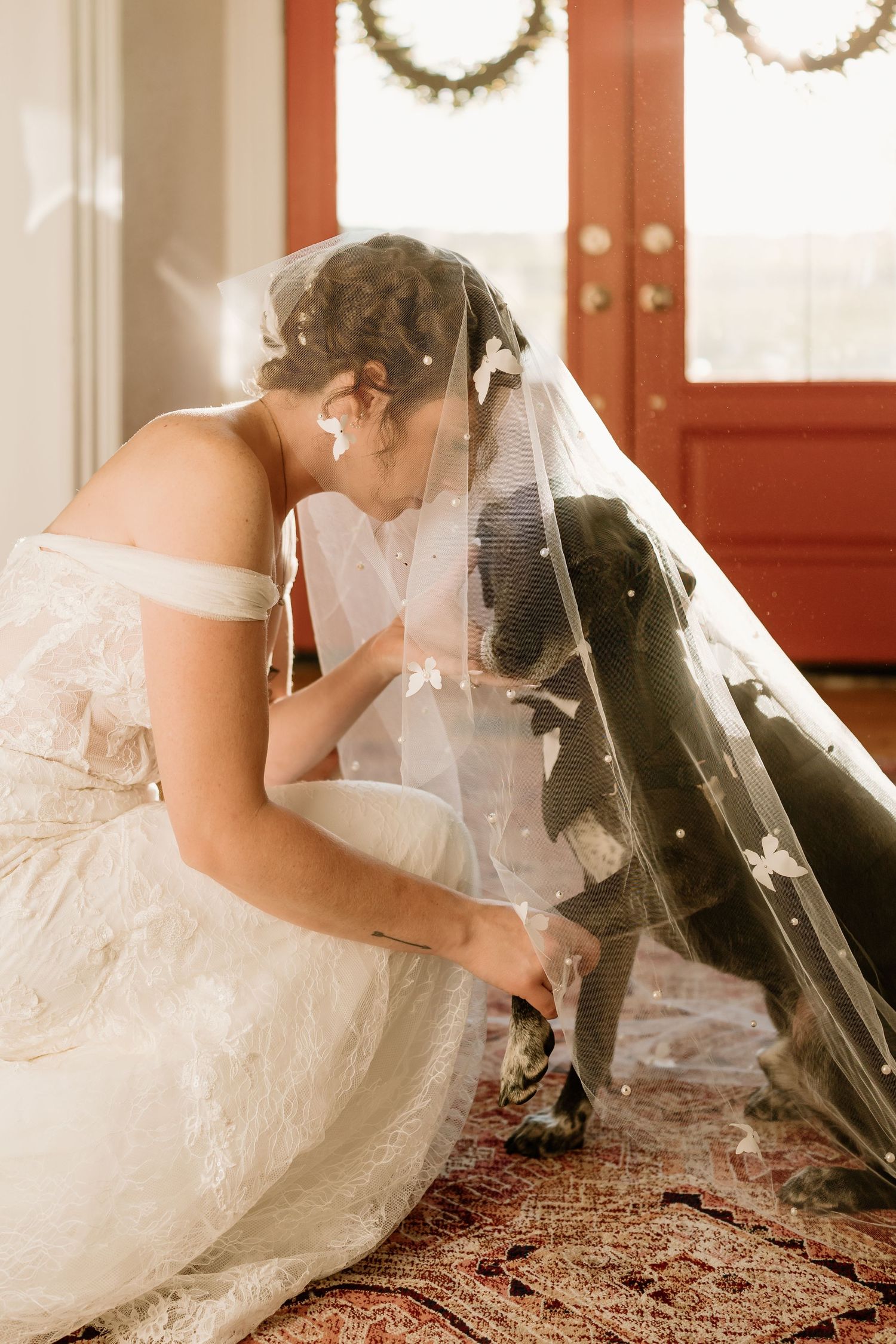 A bride in an off-shoulder wedding dress kneeling on a patterned rug by red doors.