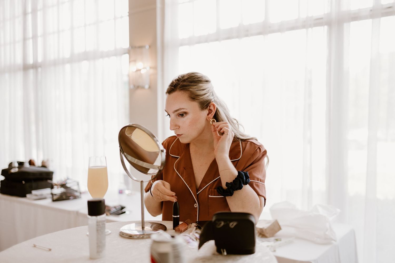 Person in silk robe applies makeup at vanity table with natural window lighting.