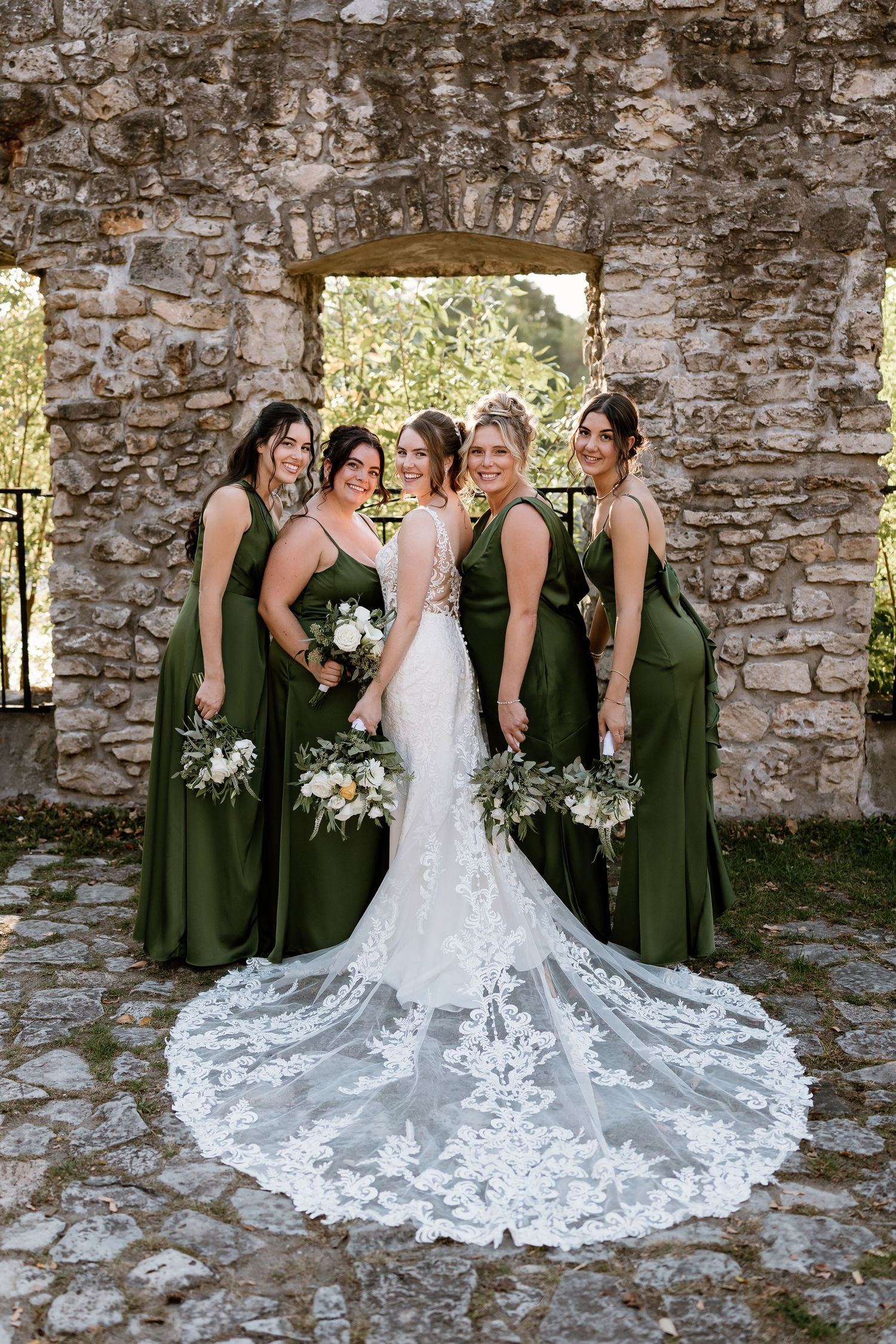 Wedding party in olive green dresses and white gown pose against rustic stone wall.