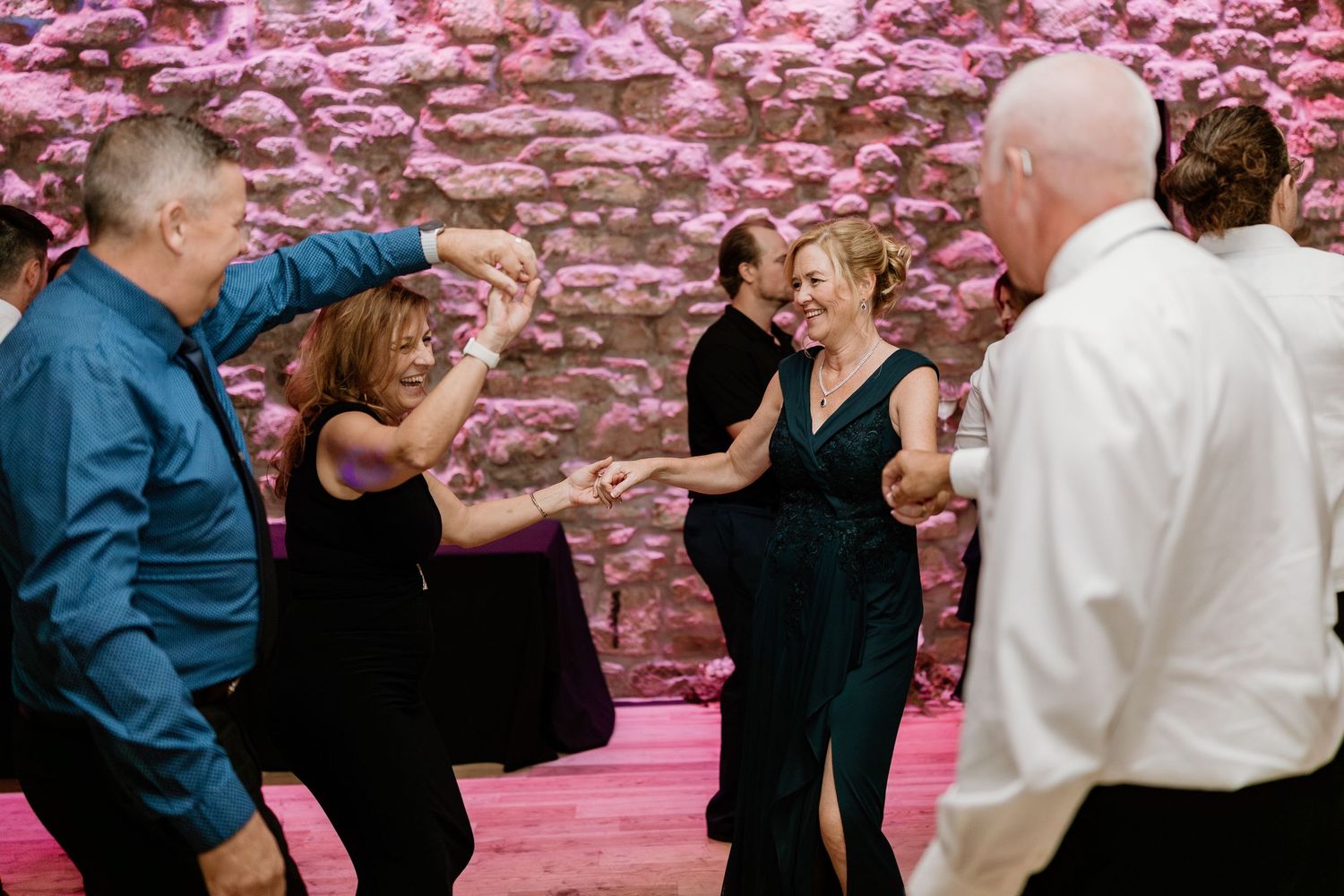 Wedding celebration with couples dancing under pink lighting against stone walls.